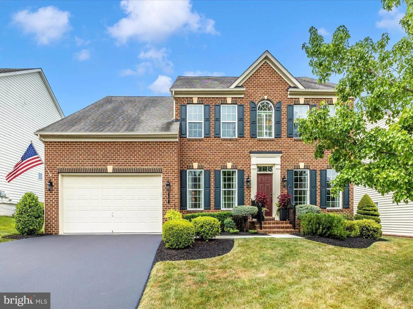 A two-story brick house with a two-car garage, black shutters, and a landscaped front yard with bushes and trees, under a blue sky with clouds.