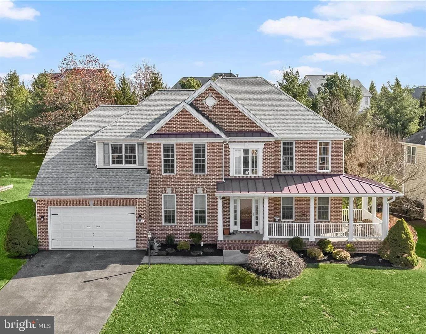 Front view of a two-story brick house with a gray shingle roof, a white garage door, front porch with white railings, and well-maintained lawn and landscaping.