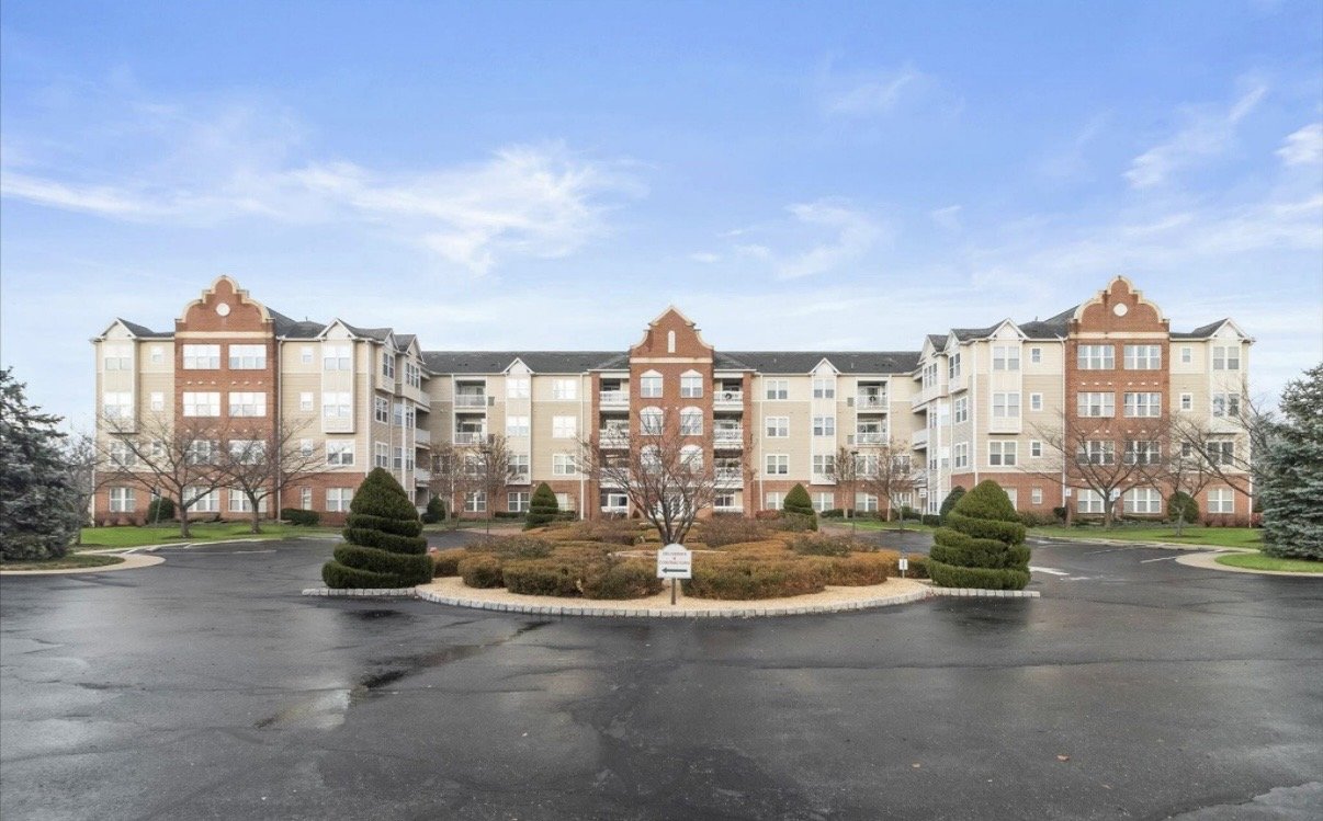 A multi-story apartment building with a manicured landscaped courtyard, leafless trees, and a paved parking area in front under a cloudy sky.