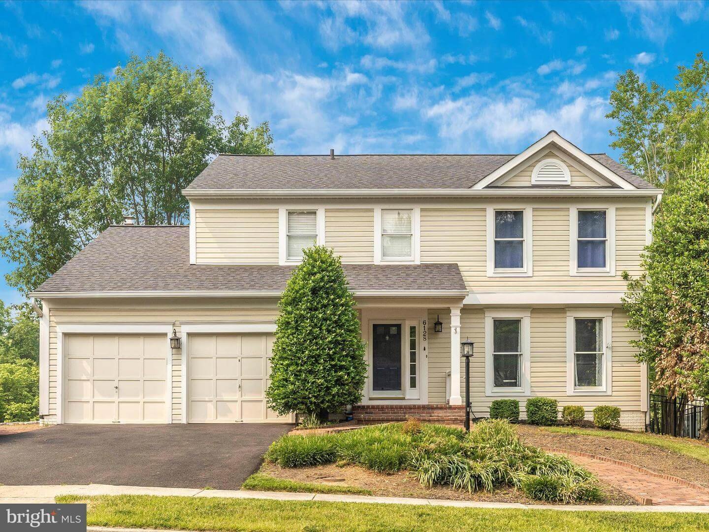Front view of a two-story suburban house with beige siding, white trim, and a three-car garage, surrounded by greenery and a well-maintained lawn under a blue sky with some clouds.
