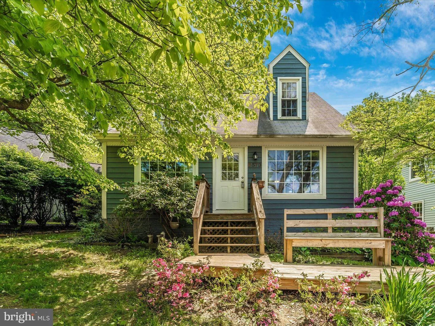 Front view of a blue house with a small porch, surrounded by green trees and flowering bushes, under a partly cloudy sky.