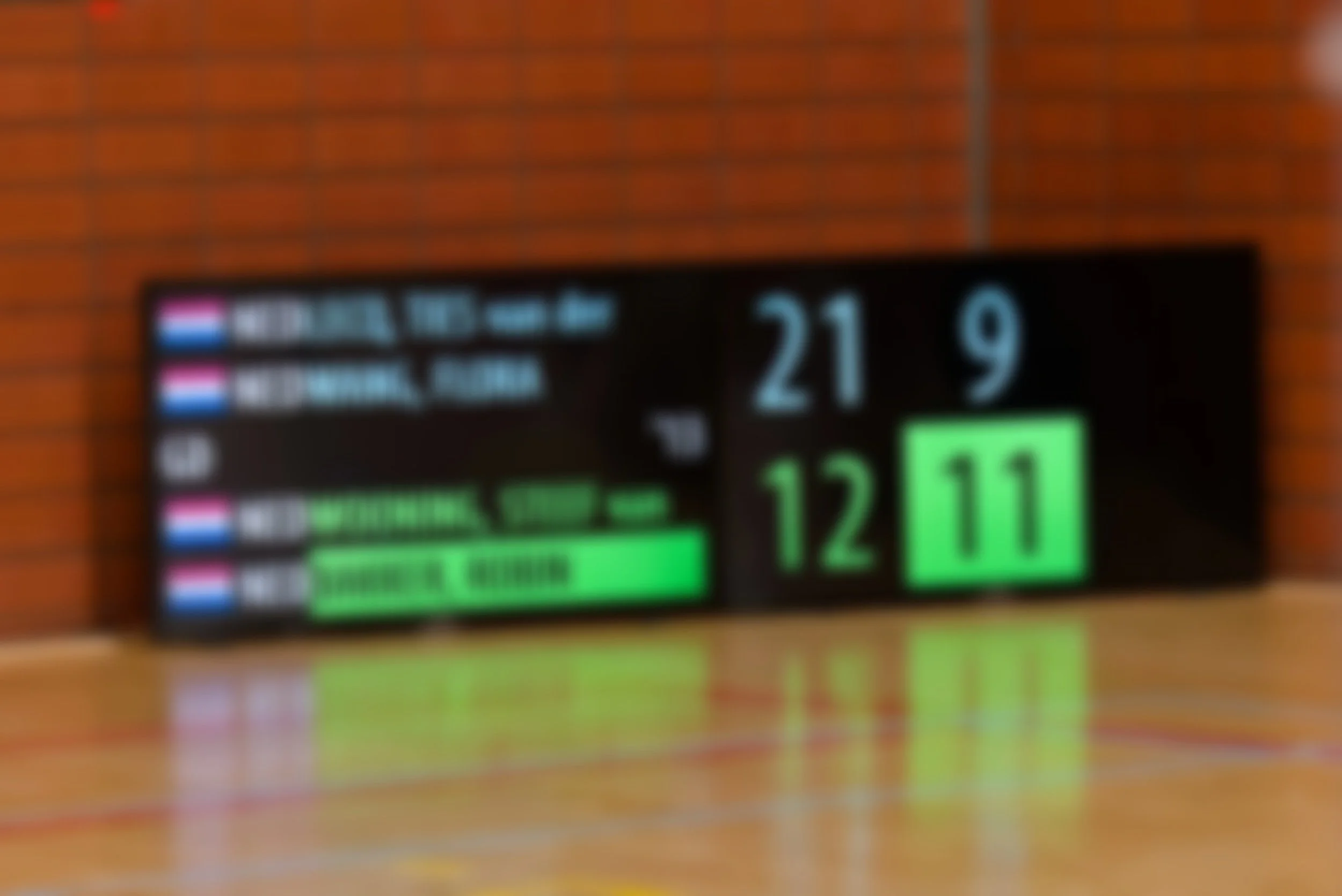 Een elektronisch scorebord met een donkere achtergrond, vertoont cijfers 21 en 9, en groene en roze details, tegen een bakstenen muur.