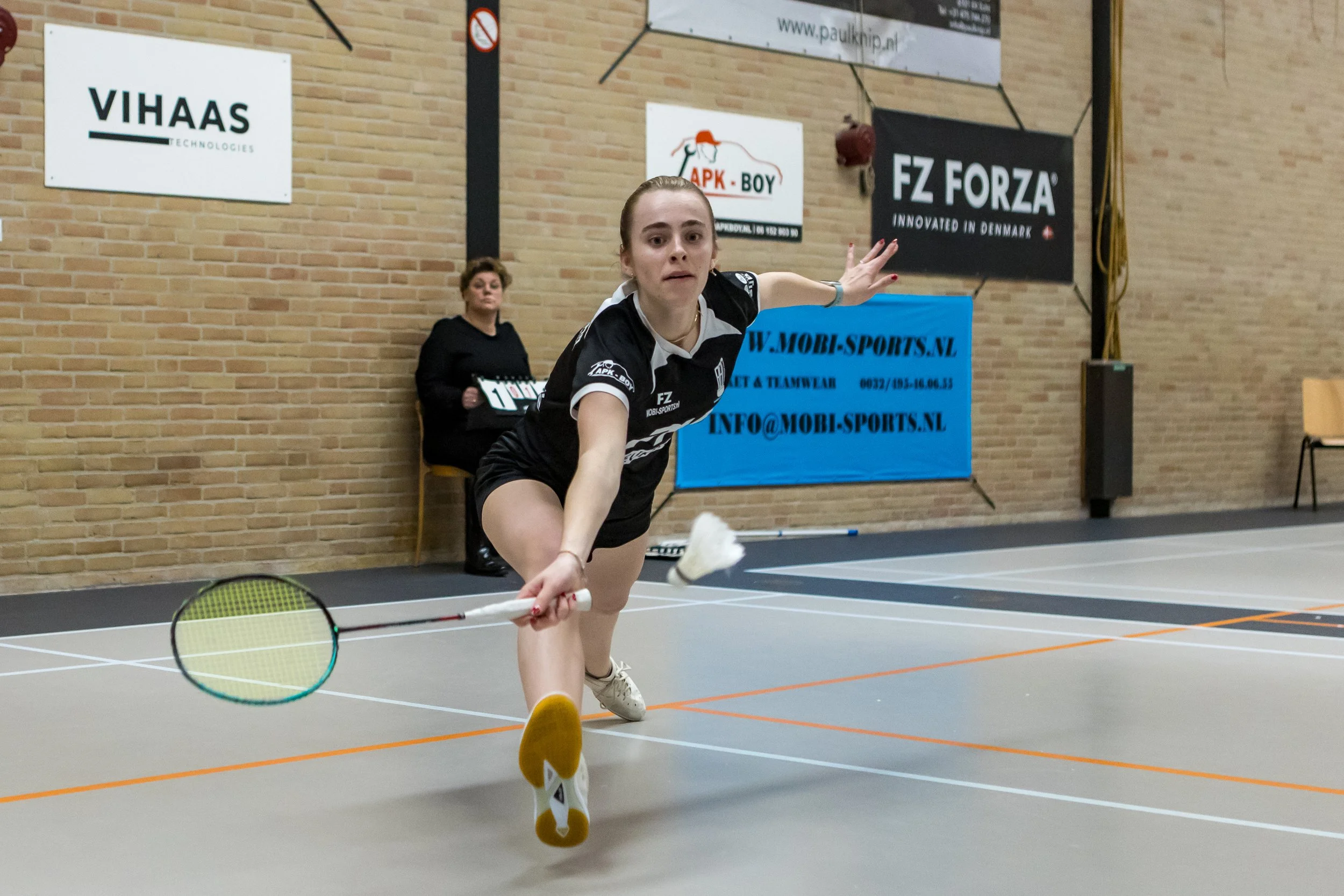 Stefanie Nijsse probeert een shuttle te slaan op een indoor sporthal, met reclameborden op de achtergrond.