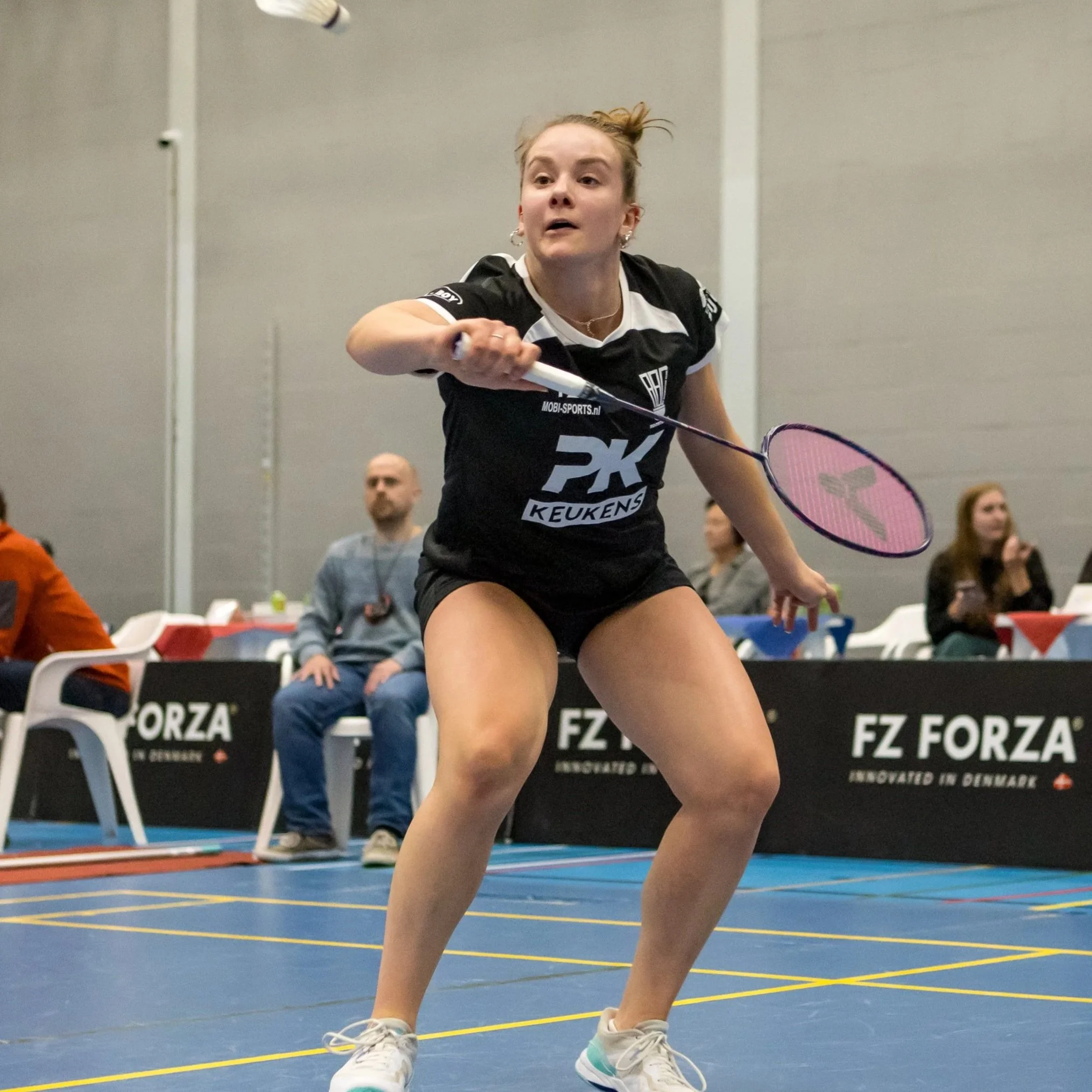 Xenia Kölmel in sportkleding die een slag uitvoert op een indoor badmintonbaan, met mensen op de achtergrond die toeschouwer of coach kunnen zijn.