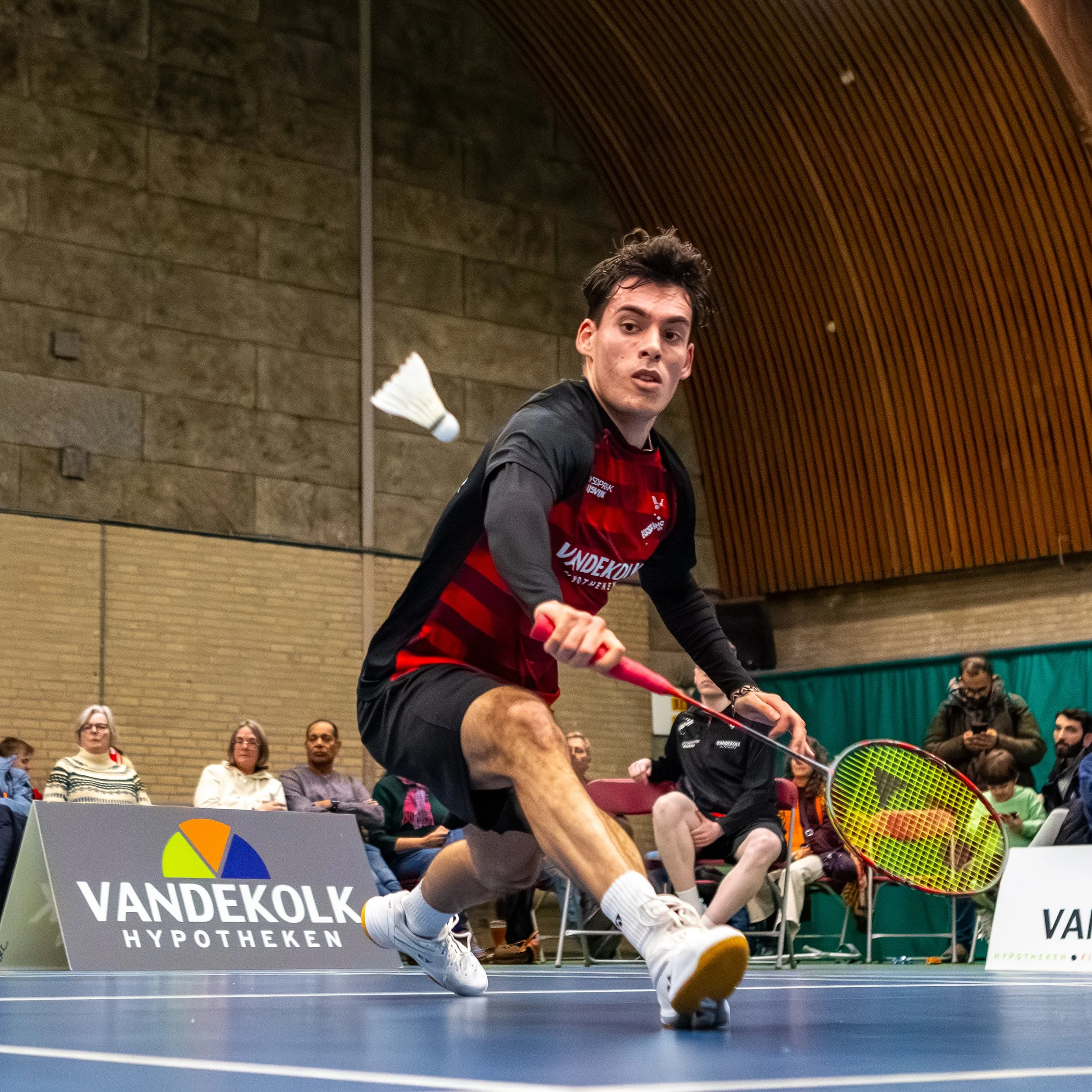 Lars Kros die een backhand slaat met een tennisracket tijdens een indoor tennistoernooi, met een publiek en reclameborden op de achtergrond.