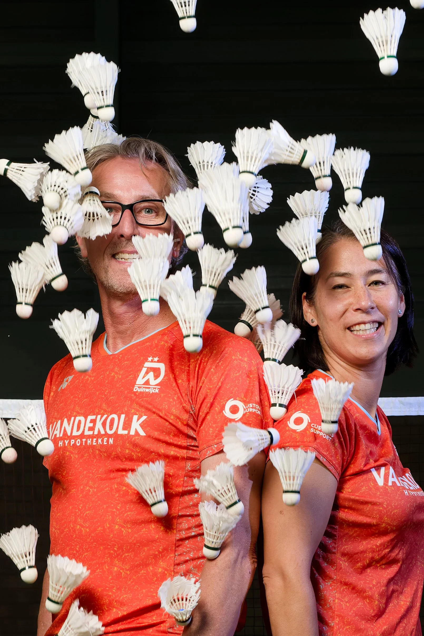 Twee badmintonspelers in oranje tenues met sponsorlogo's, omringd door vliegende shuttlebats.