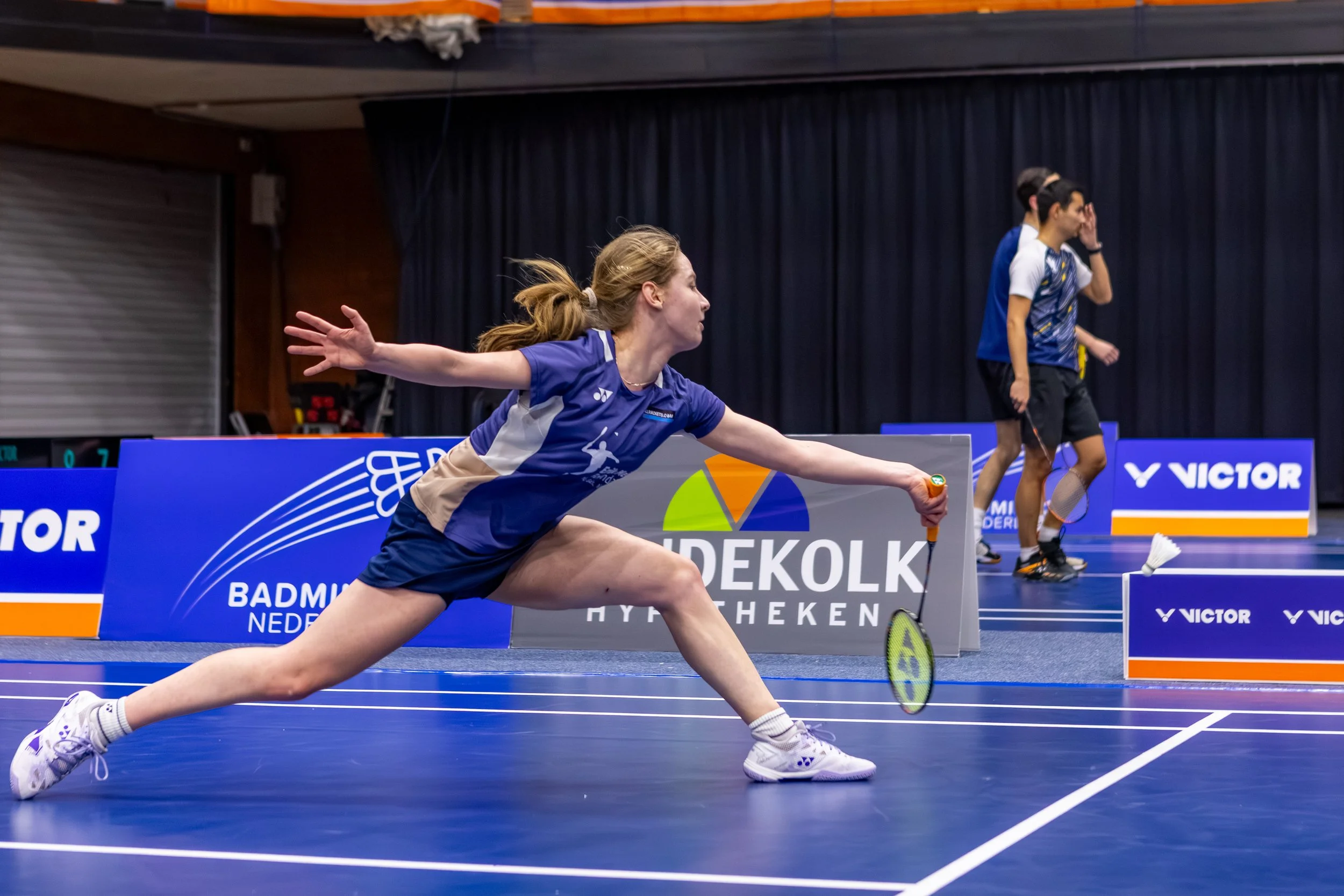 Esmeralda van Drunen in actie tijdens een wedstrijd op een blauwe baan met het logo en tekst 'Badminton Nederland' op de achtergrond.