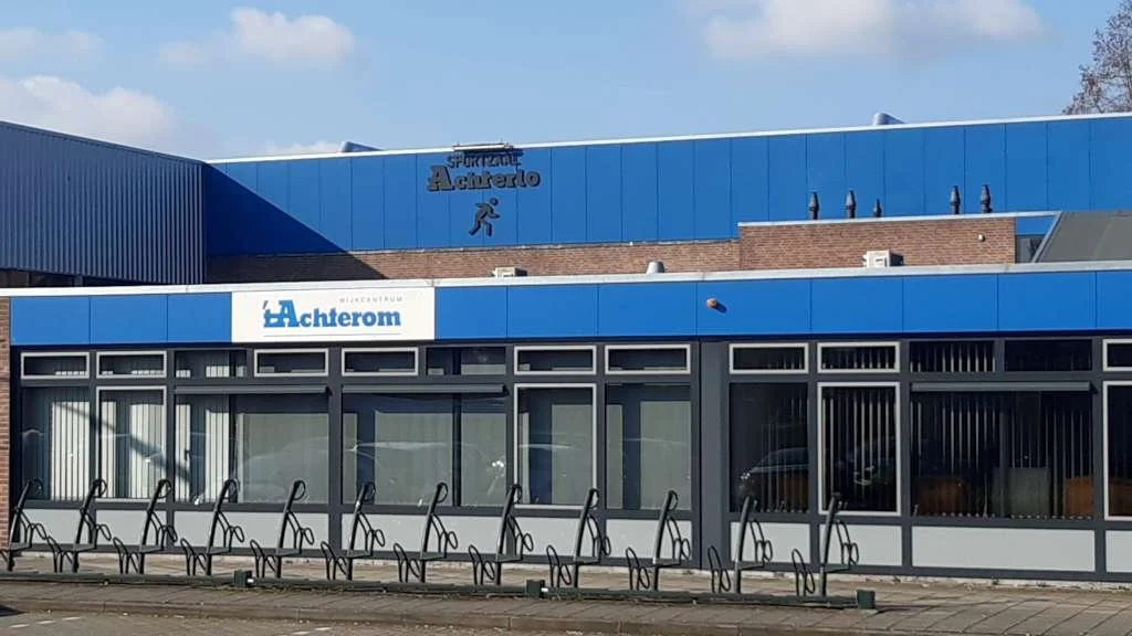 Een winkel met grote ramen, fietsenrekken voor de winkel en een blauw bord met de tekst 'Achterom', onder een blauw gedeelte van het gebouw met een sport afbeelding en de tekst 'Sportcentrum Achterom'.