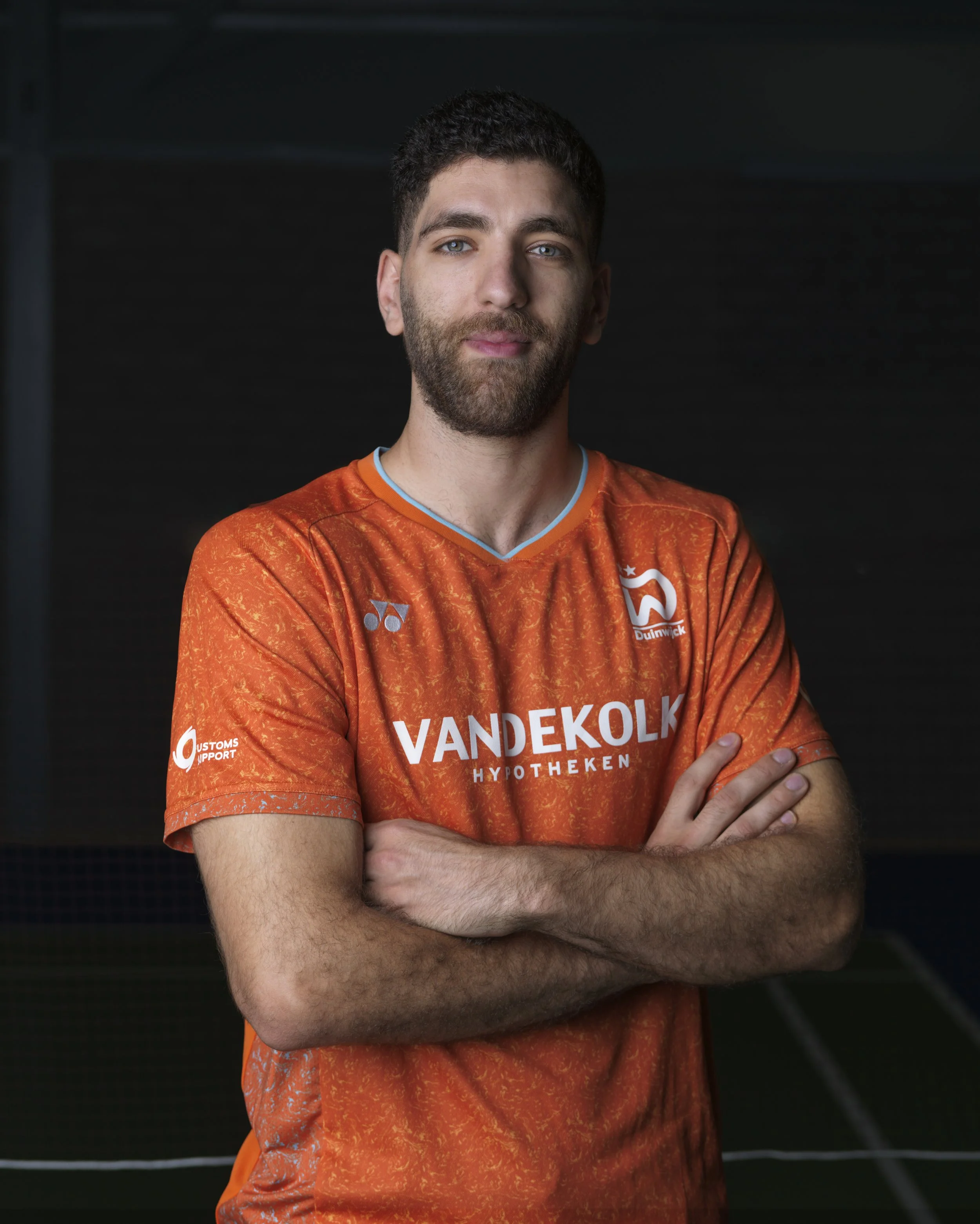 Portret van een jonge man met een donkere achtergrond, gekleed in een oranje sportshirt met sponsorlogo's.