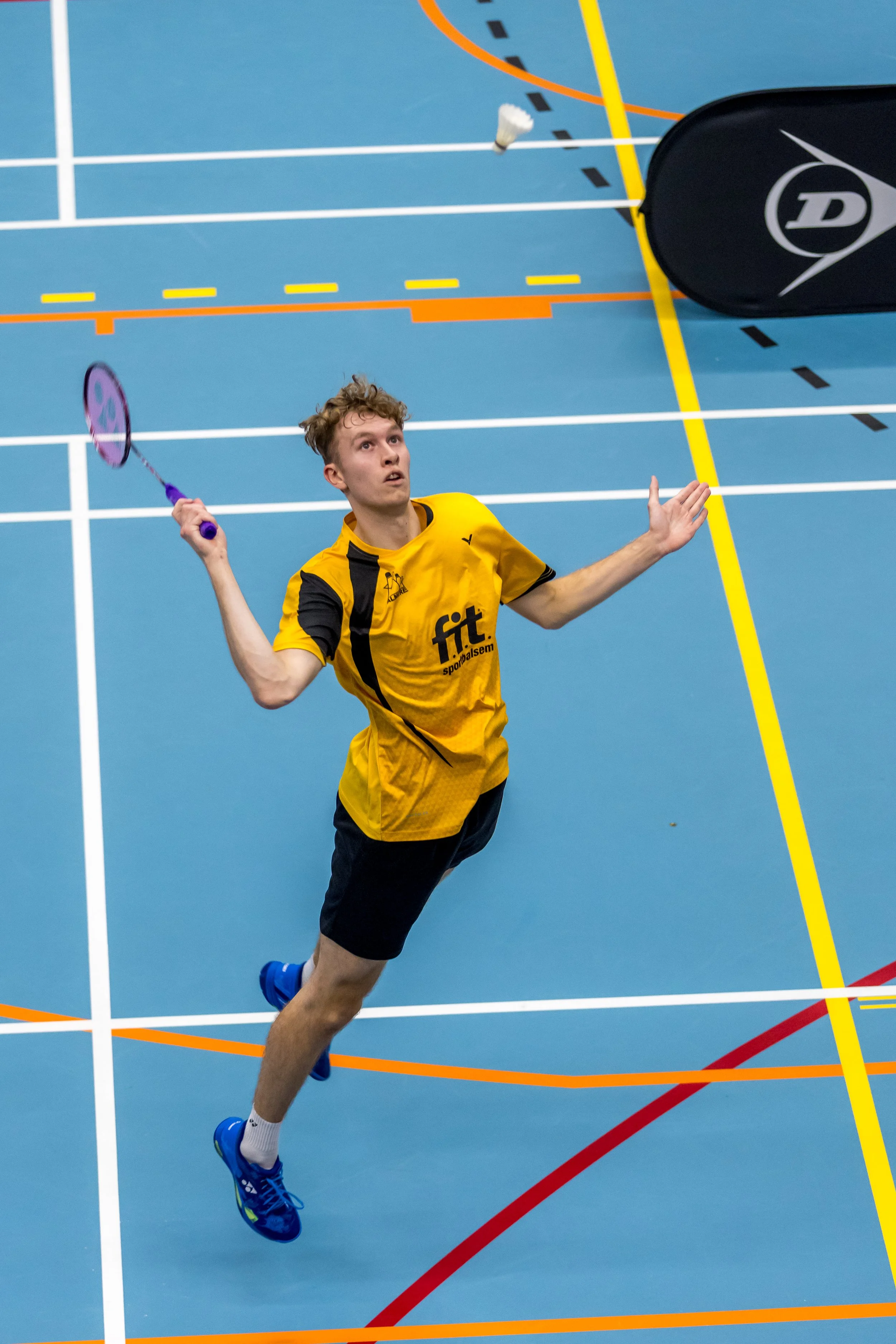 Dennis Koppen in een gele sportoutfit die badminton speelt op een blauwe indoorbaan, in beweging met een badmintonracket in de hand.
