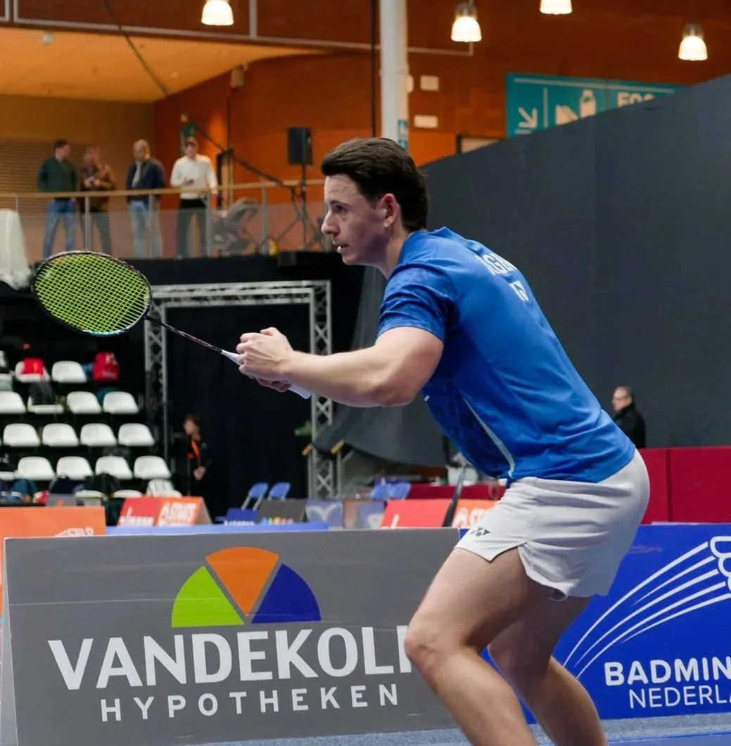Een man die badminton speelt in een sporthal, gekleed in een blauwe shirt en witte shorts, klaar om de shuttle te slaan.
