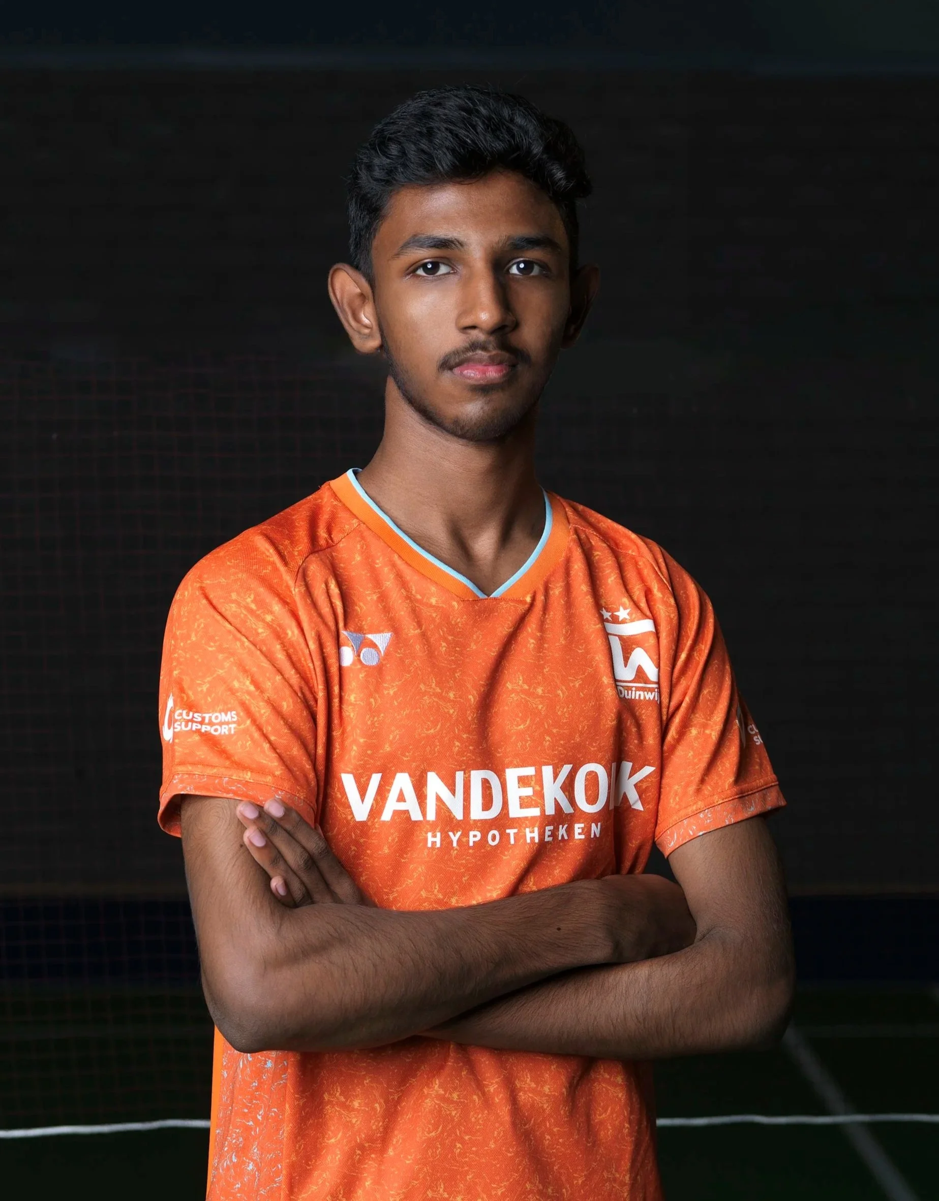 Portret van een jonge man met korte, donkere haar, gekleed in een oranje sporttenue met sponsorlogo's, staande voor een donkere achtergrond.