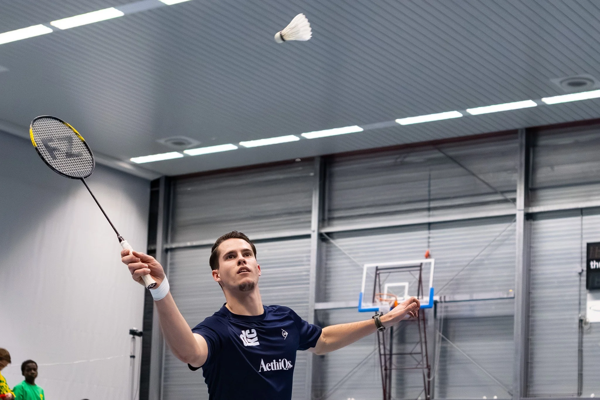 Gijs Duijs die badminton speelt in de hal van BC DKC met een shuttle en een racket in zijn hand, en andere mensen op de achtergrond.