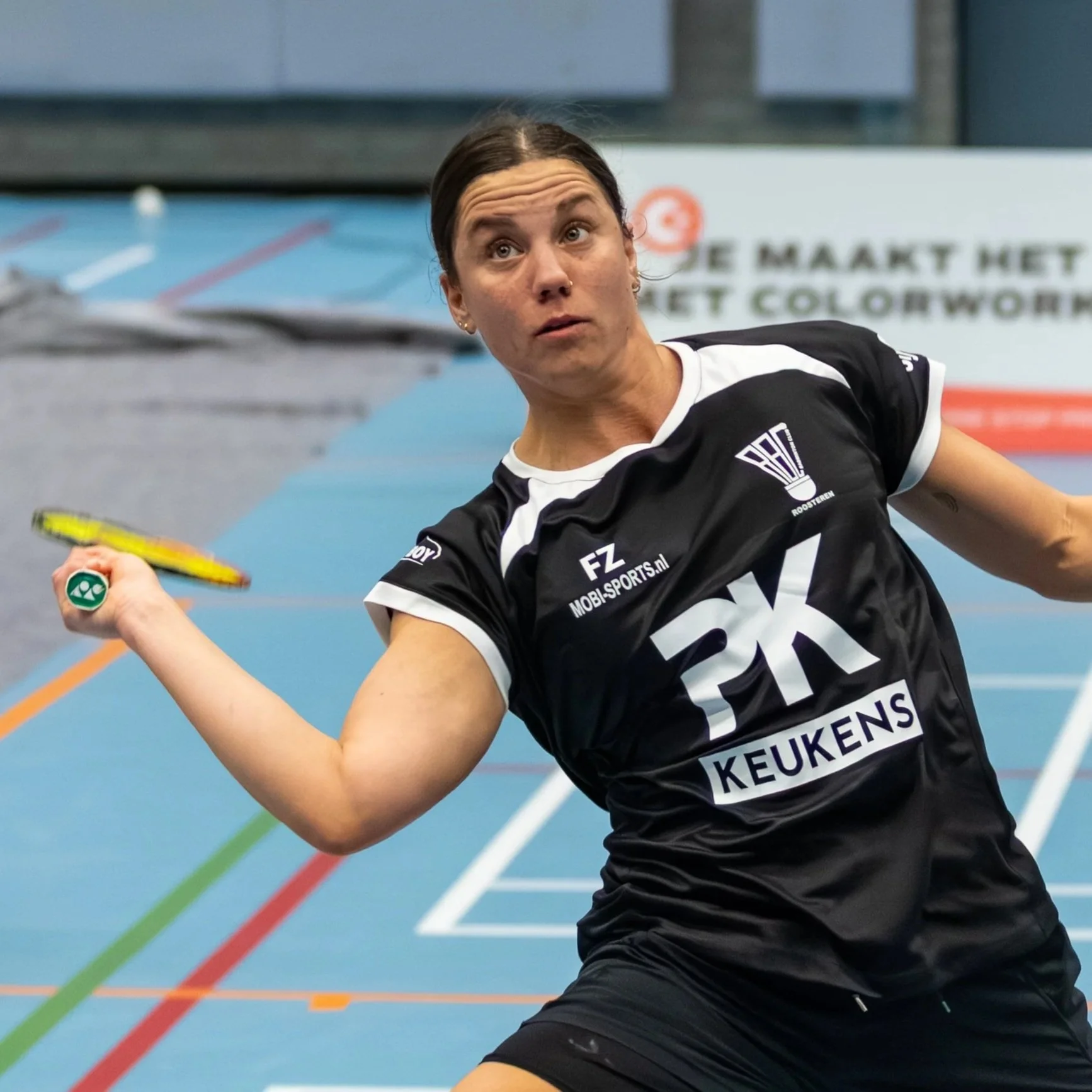 Flore Vandenhoucke met lang, donker haar die badminton speelt op een indoorbaan met meerdere gekleurde lijnen.
