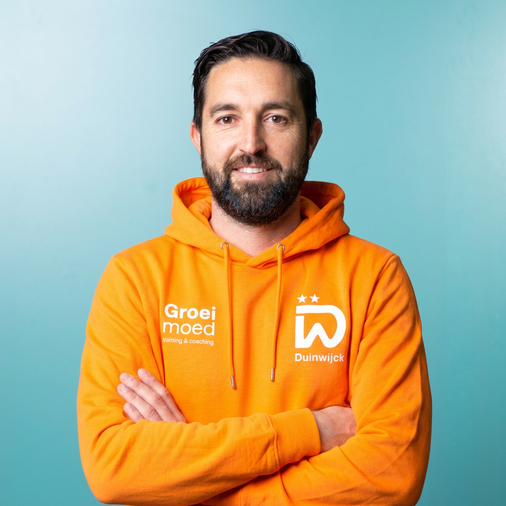 Man in een oranje sportshirt met armen over elkaar voor een donkere achtergrond.