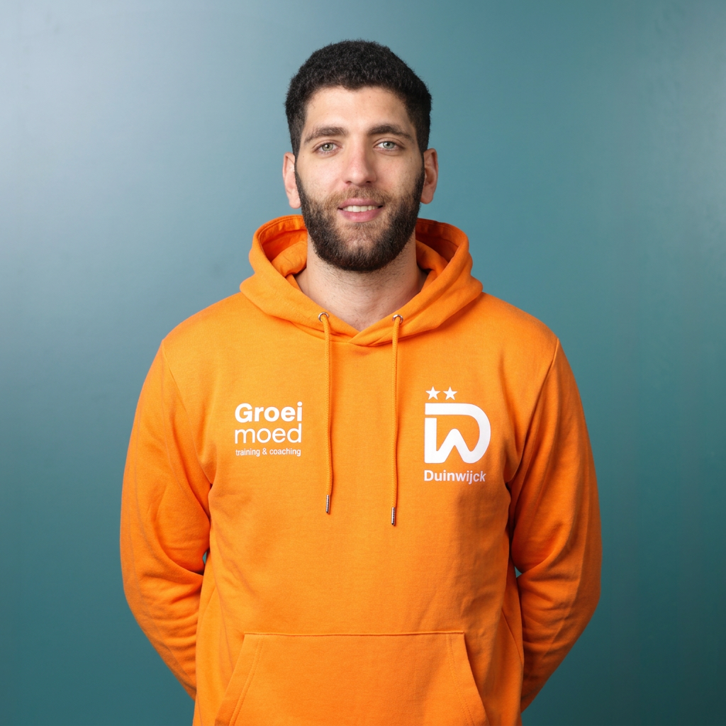 Portret van een jonge man met blauwe ogen en getrimd haar, gekleed in een oranje sport T-shirt met witte en blauwe details, staand tegen een zwarte achtergrond.