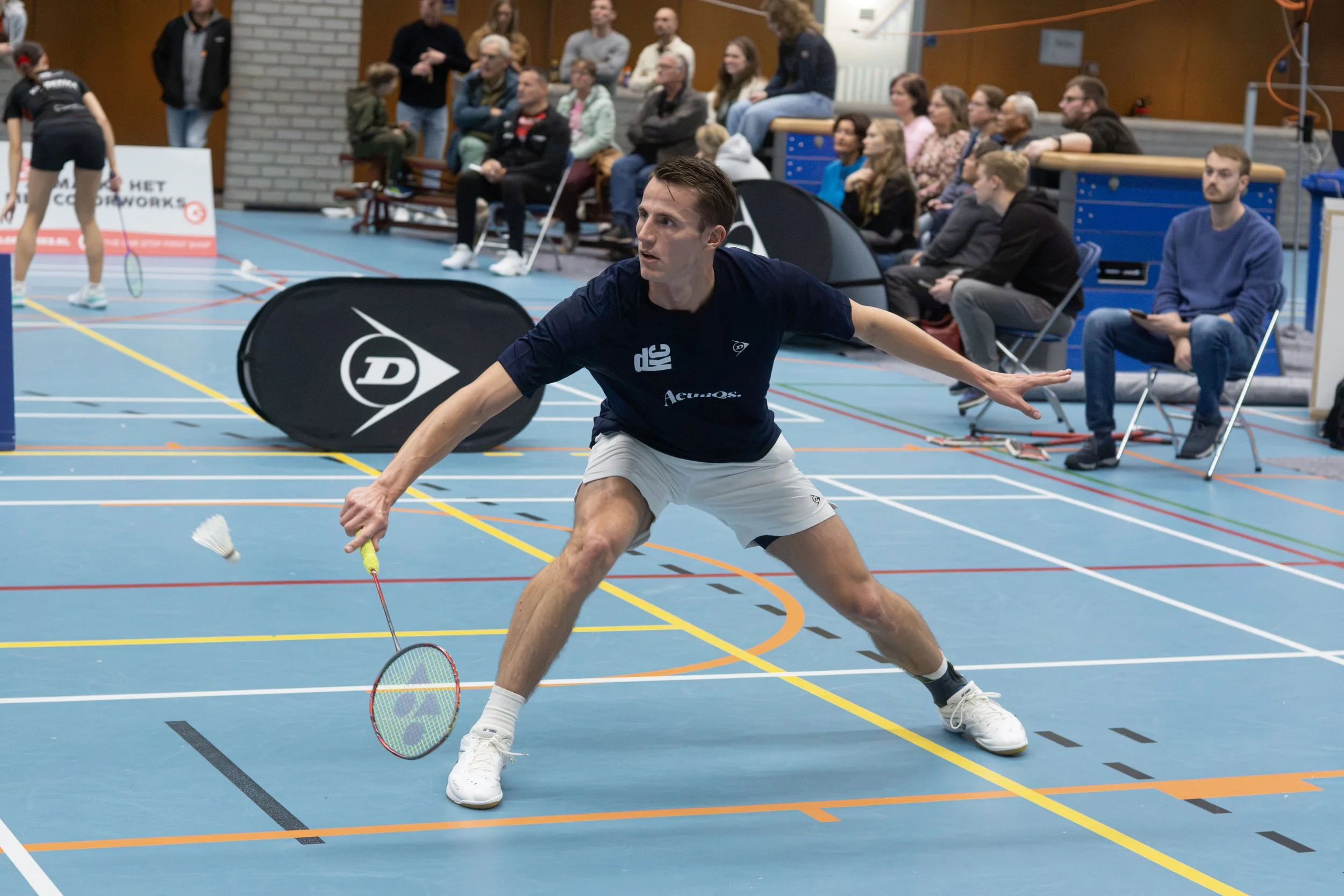Een man speelt badminton in een sporthal, met een volle tribune en twee vrouwen op de achtergrond. Naast hem ligt een zwarte balpak met een logo erop.
