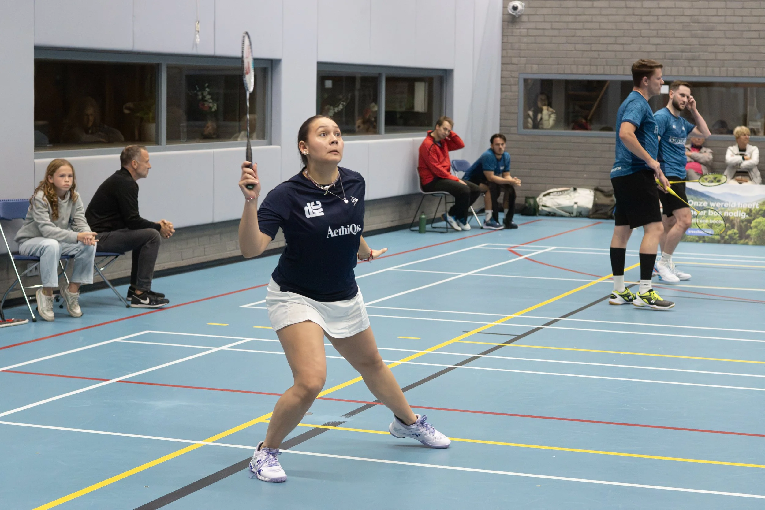 Een vrouw speelt badminton in een binnenhal, omringd door enkele toeschouwers en andere spelers die wachten.