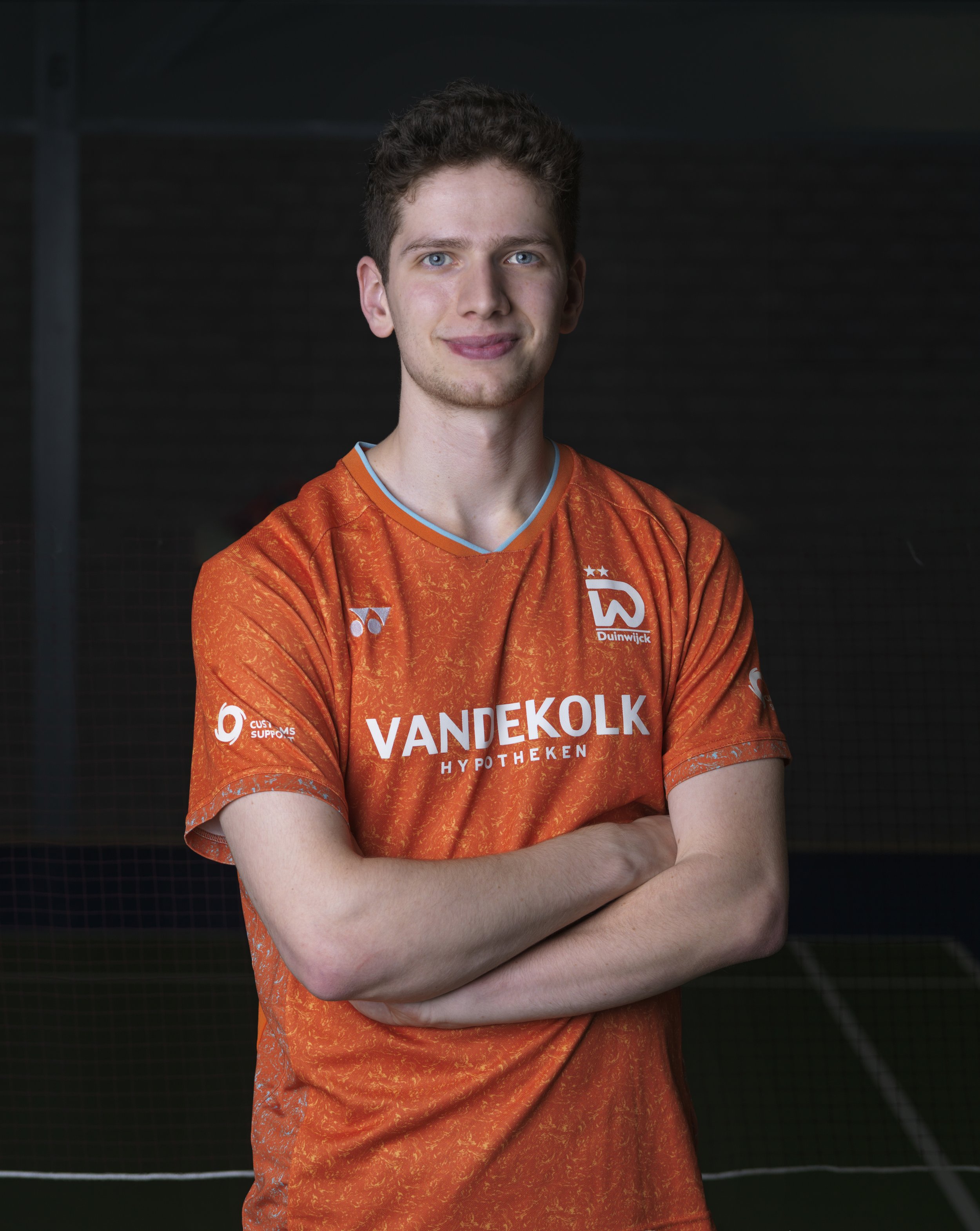 Een jonge man met blauwe ogen en kort, krullend haar, gekleed in een oranjerood sportshirt met tekst en logo's, staand met gekruiste armen voor een donkere achtergrond.