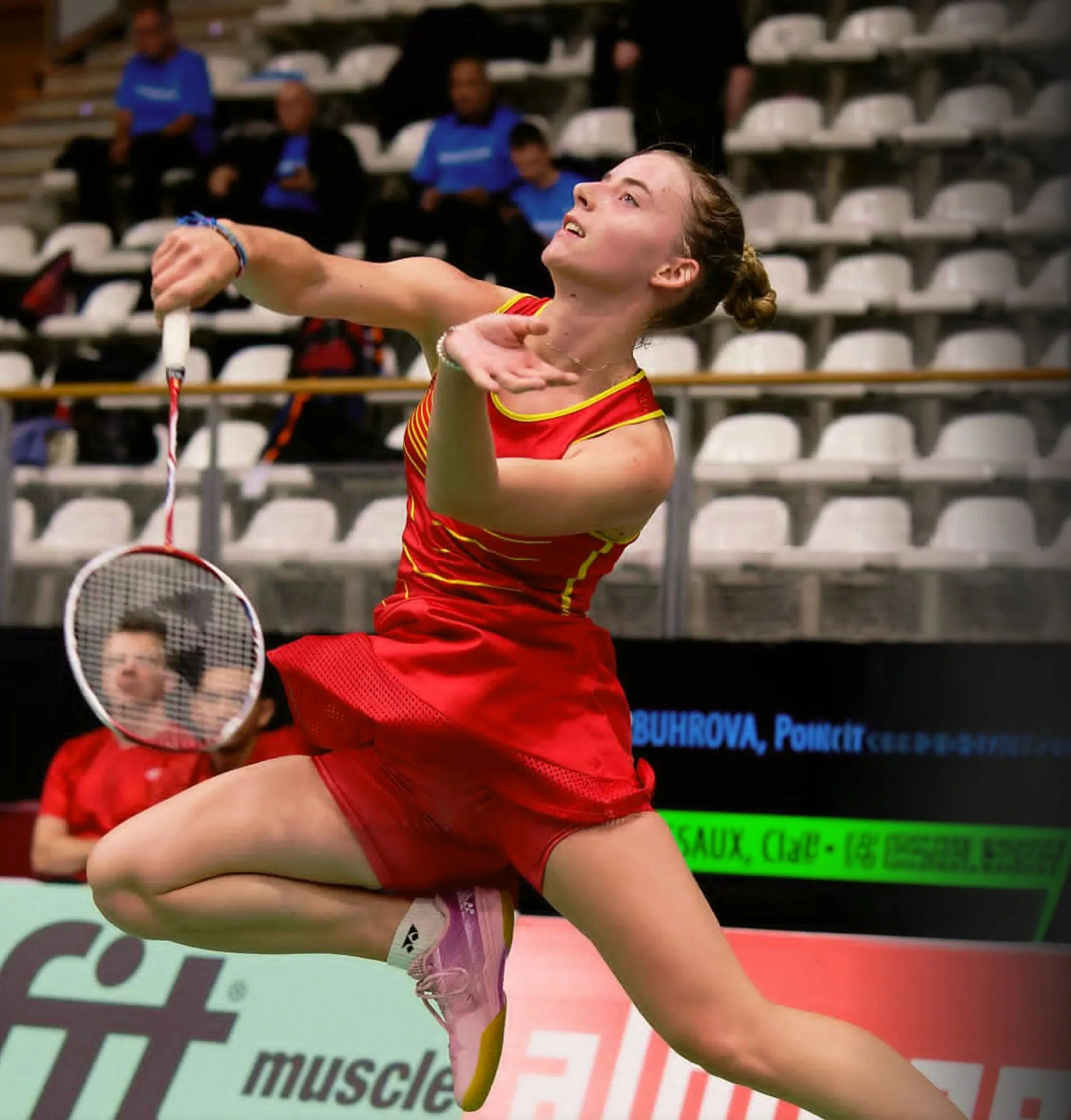 Een vrouwelijke badmintonspeler in een rode outfit, klaar om te slaan met haar racket, terwijl haar tegenstander in de achtergrond zich voorbereidt op de volgende slag, op een indoor sporthal met meerdere lege zitplaatsen.