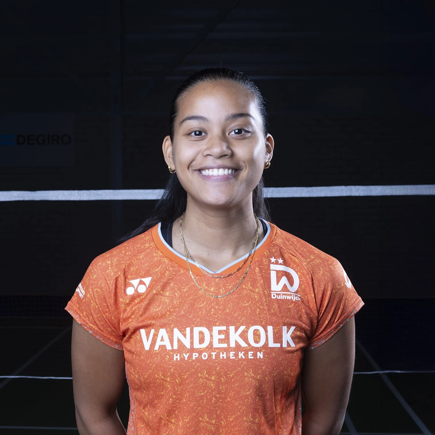 Een jonge vrouw in een oranje sportshirt met sponsorlogo's, lachend voor een donkerbruine achtergrond.