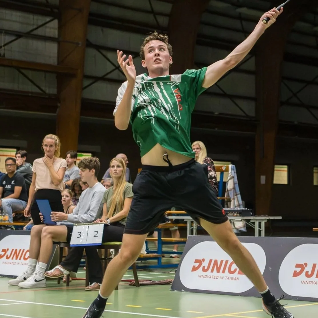 Dion Rovers die badminton speelt op een indoor sporthal, met een publiek dat toekijkt op de achtergrond.