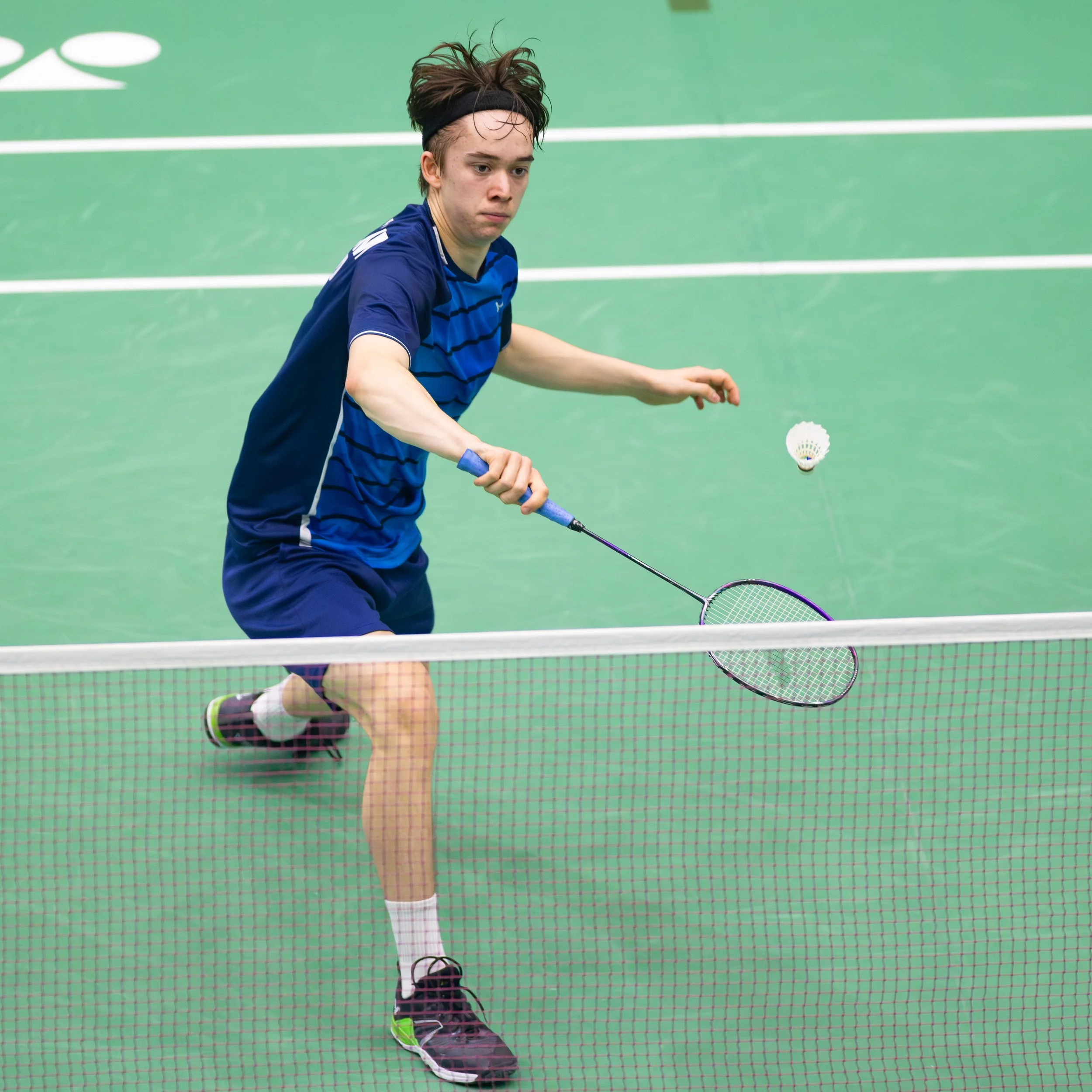 Angus Meldrum in een blauw T-shirt en donkergroene schoenen speelt badminton op een groene indoor baan, die wordt gezien door een net.