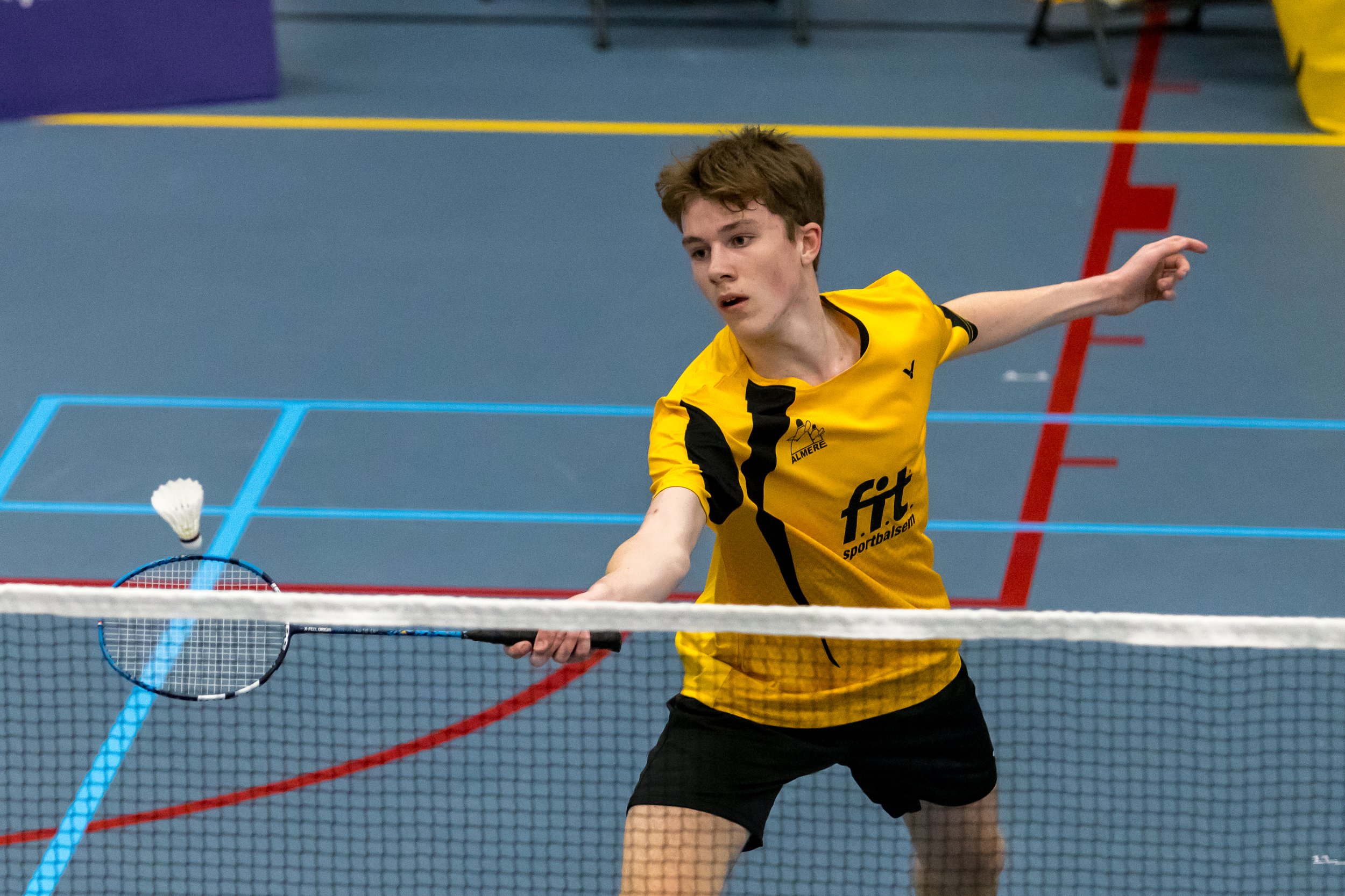 Joep Strooper speelt badminton in een sporthal, hij draagt een gele sportoutfit en houdt een badmintonracket vast terwijl hij zich uitstrekt.