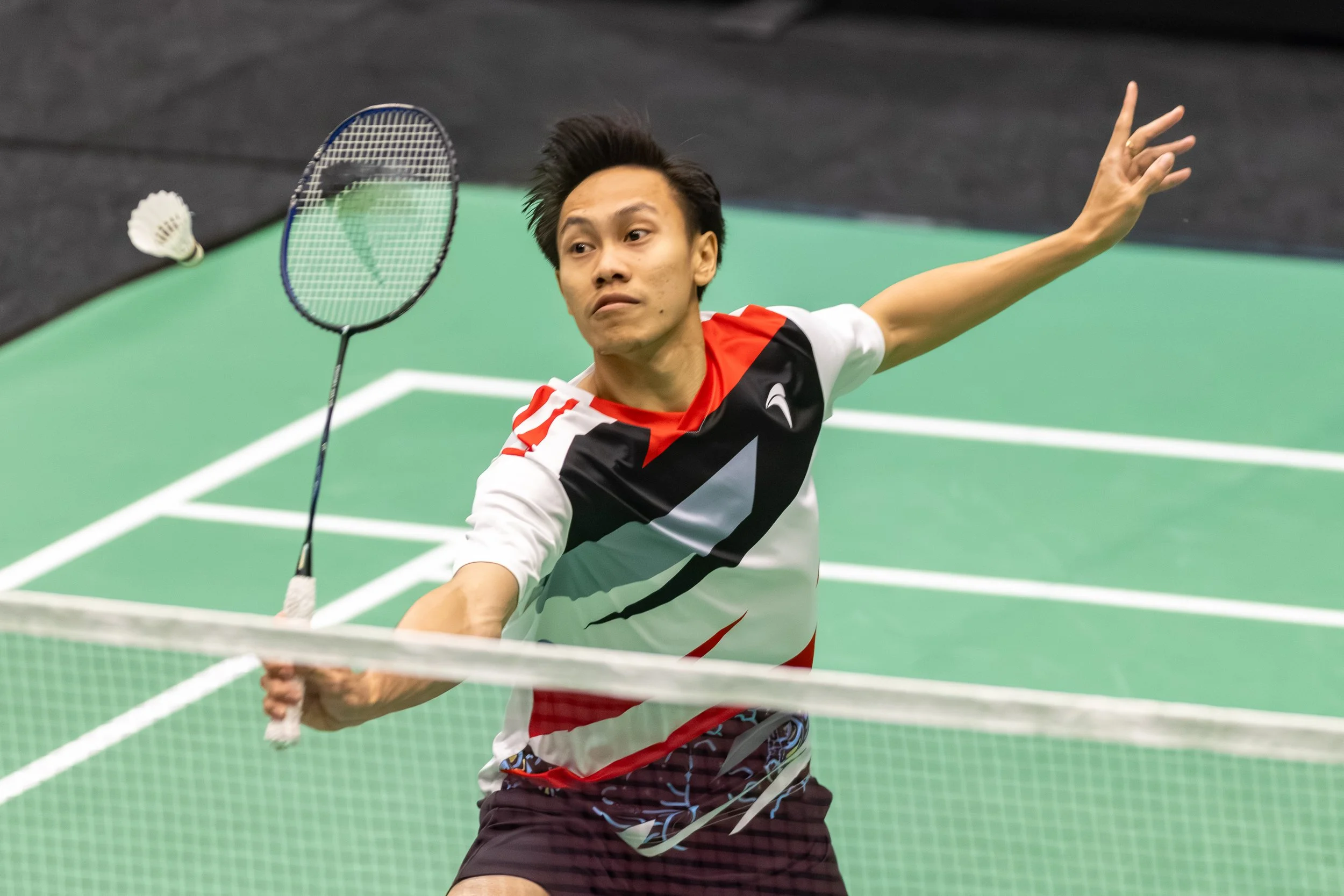 Ade Resky Dwicahyo die badminton speelt op een groene sportvloer, hij is bezig met een slag terwijl hij zich uitstrekt.