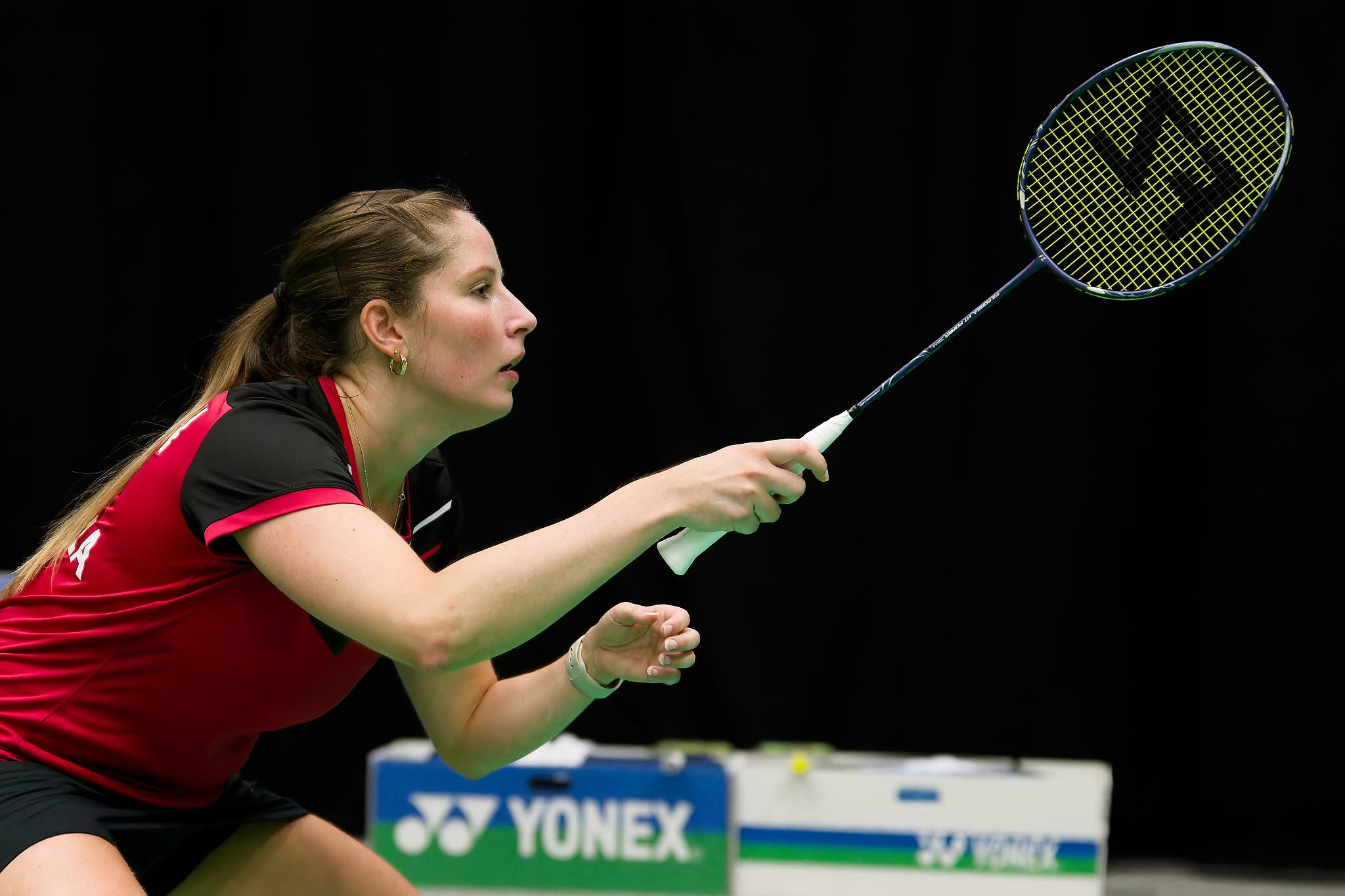 Iben Bergstein in actie, klaar om een shuttle te slaan, met een donkere achtergrond en een Yonex-bord op de baan.