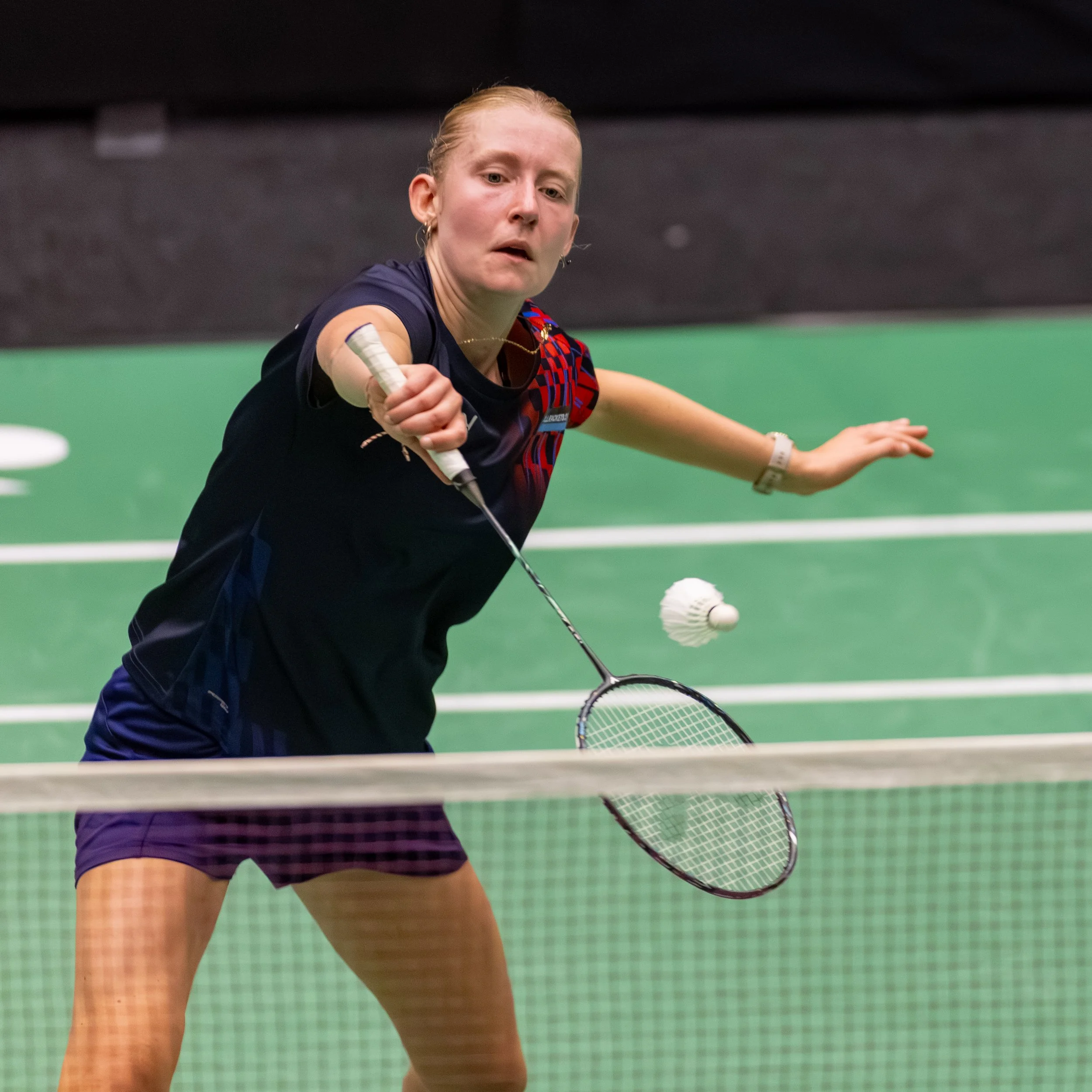 Veda Diemel, een vrouwelijke badmintonspeelster slaat de shuttle met haar racket tijdens een wedstrijd op een groene badmintonbaan.