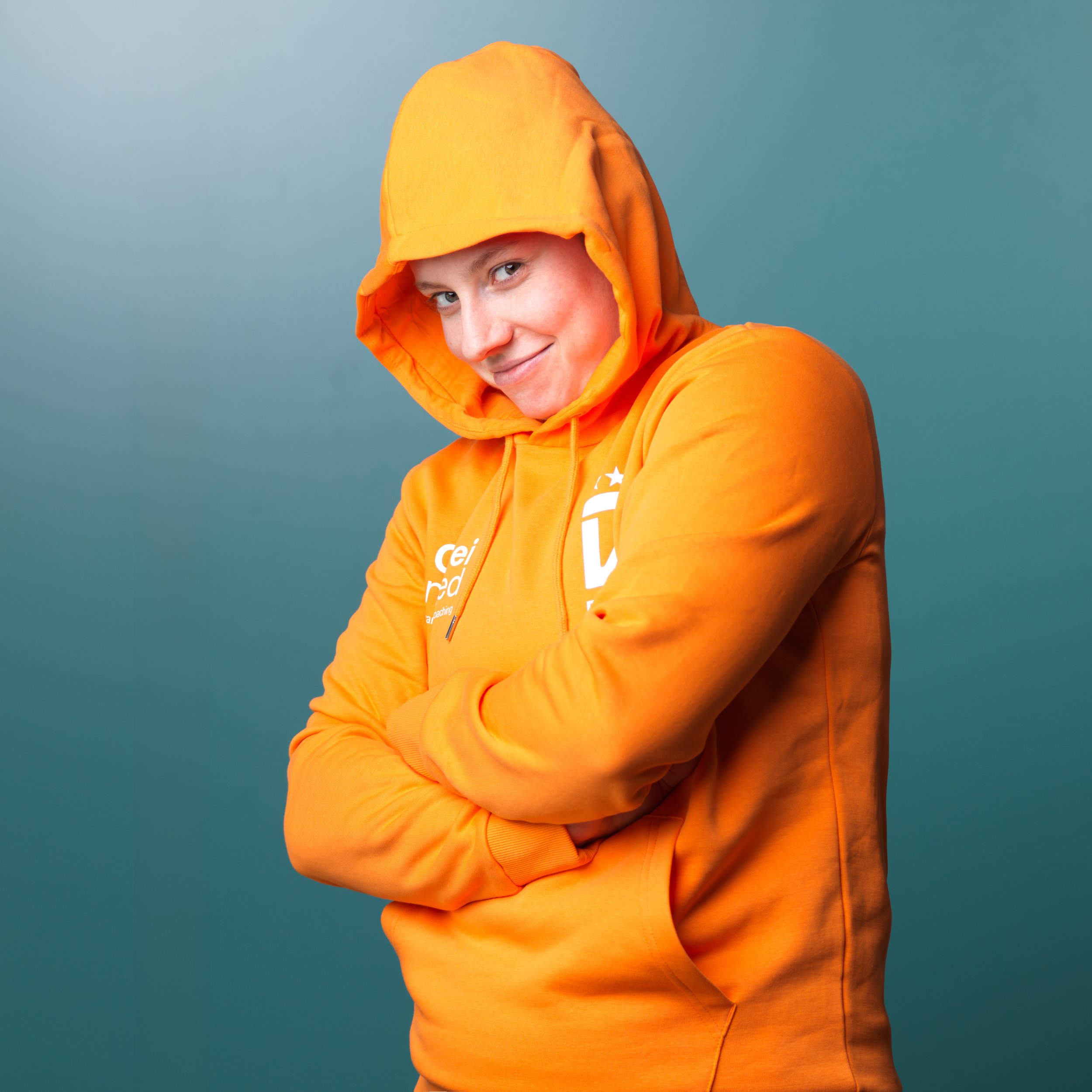 Close-up van een glimlachende jonge vrouw met rood haar, in een oranje sportshirt met tekst en logo's, voor een donkere achtergrond.