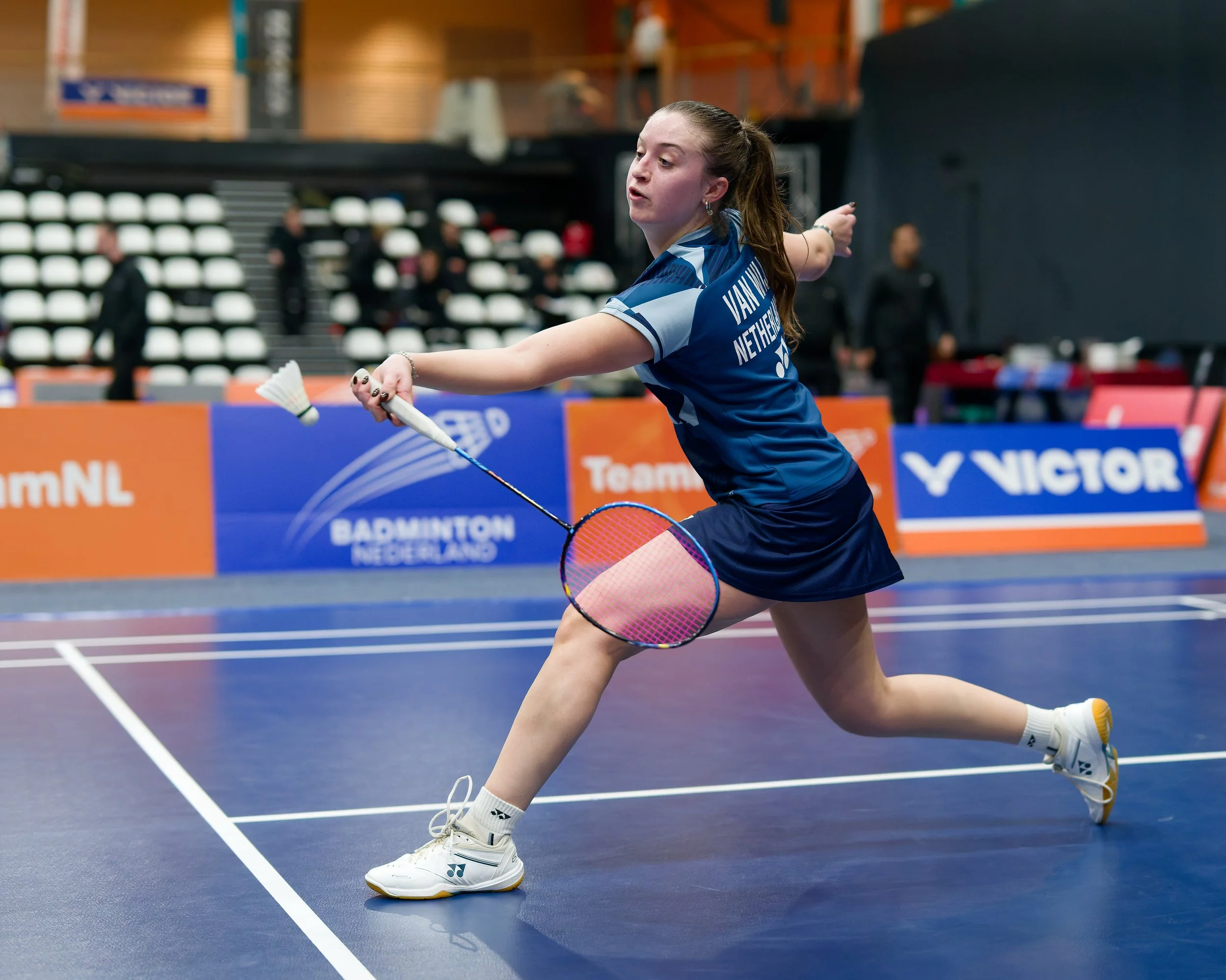 Anouk van Wijk, een vrouwelijke badmintonspeler in een blauwe uitrusting speelt op een indoor baan, haar racquet klaar om de shuttle te slaan.