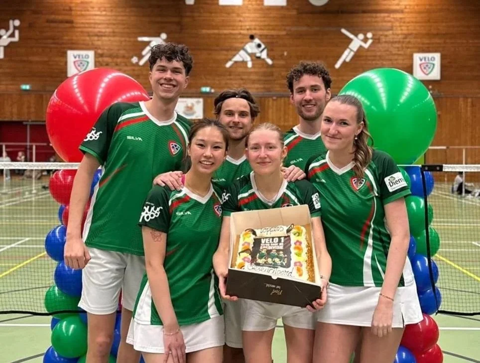 Vrouwelijk badmintonteam in groene sportkleding poseert in een sporthal, met een taart en ballonnen.