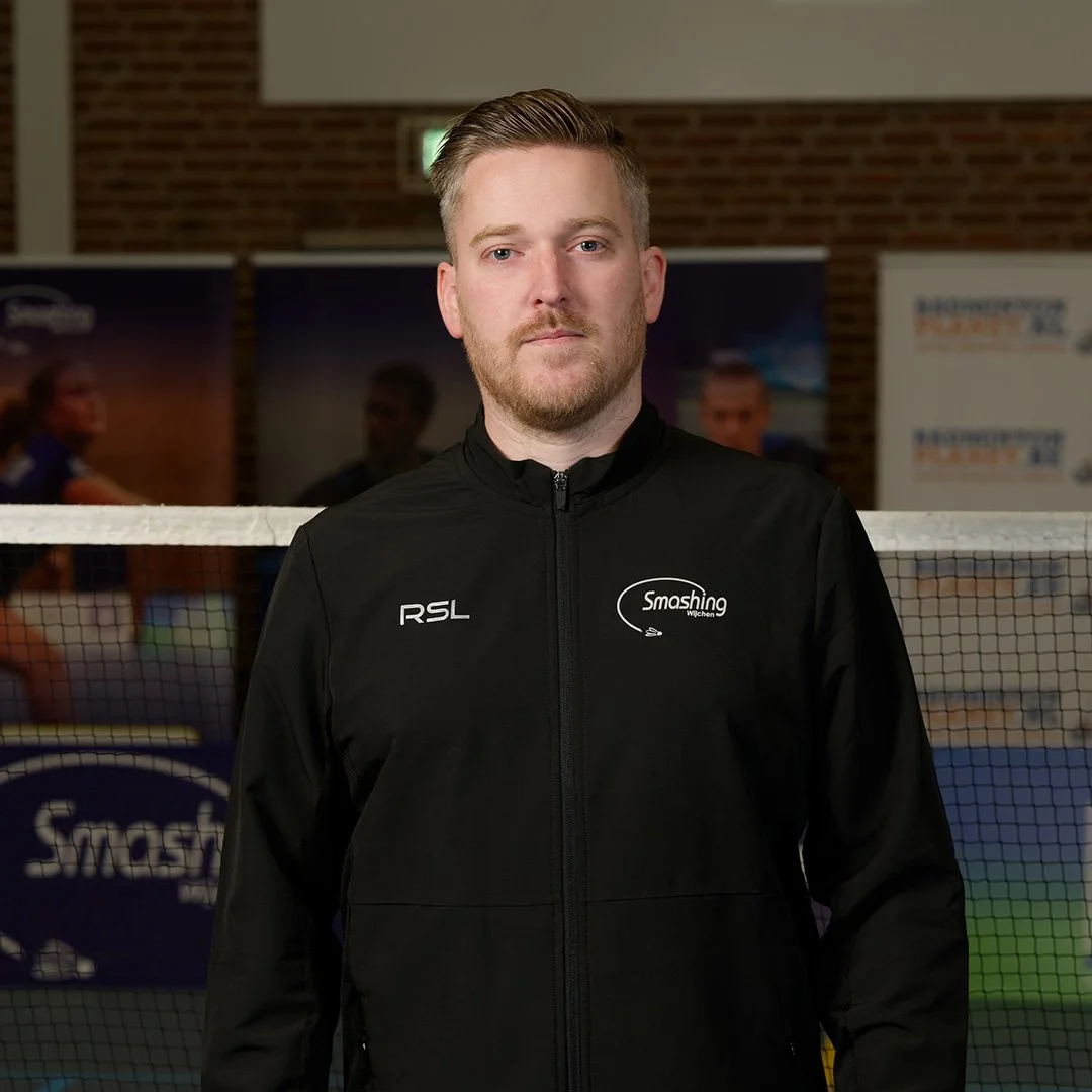 Man in zwart sportjack met logo's voor een badmintonvereniging, standing voor een badminton net in een sporthal.