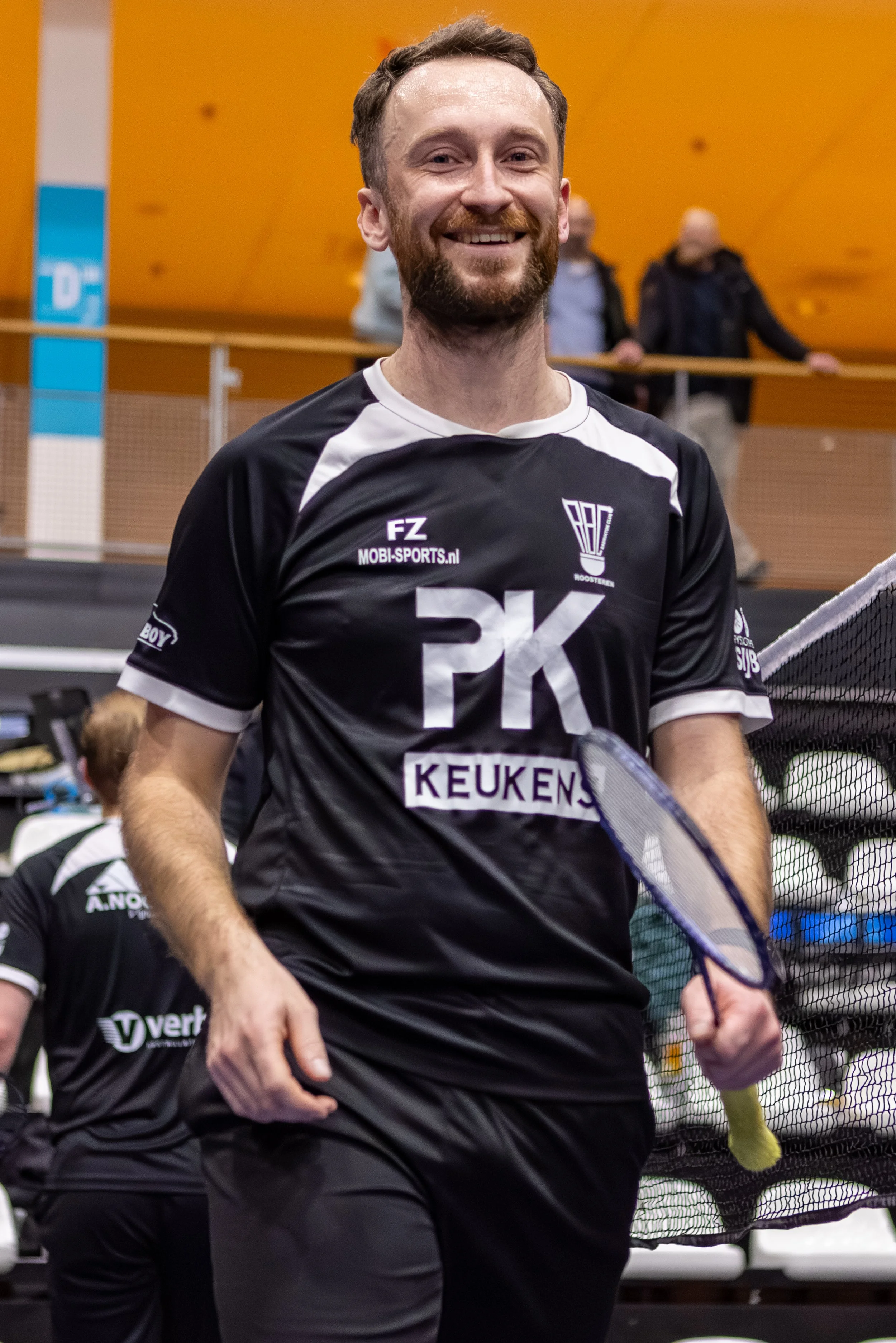 Aan de voorgrond een lachende man met een baard in een zwart en wit sportshirt met sponsors. Op de achtergrond mensen in een sporthal.