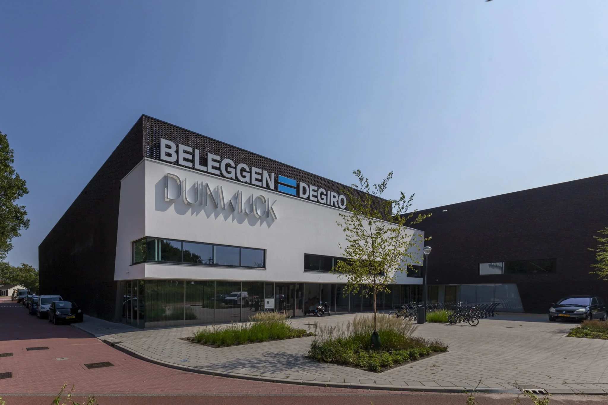 Moderne kantoorgebouw met grote ramen en een bovenlicht, met parkeerplaatsen en fietsen voor het gebouw, onder een heldere blauwe hemel.