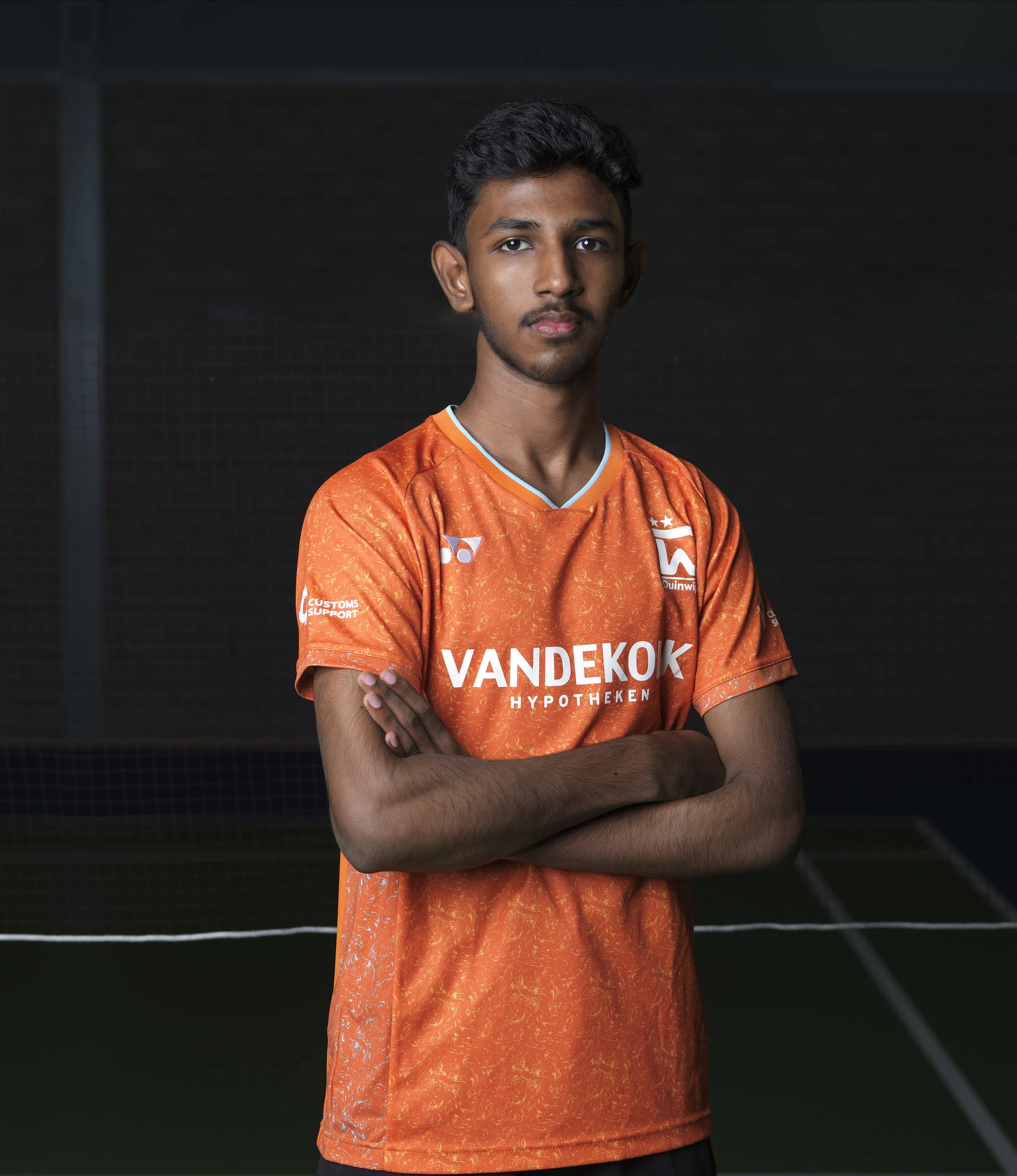 Tiener jongetje met kort zwart haar en een sportshirt met oranje en witte kleuren, staand met gekruiste armen voor een tennisbaan achtergrond.