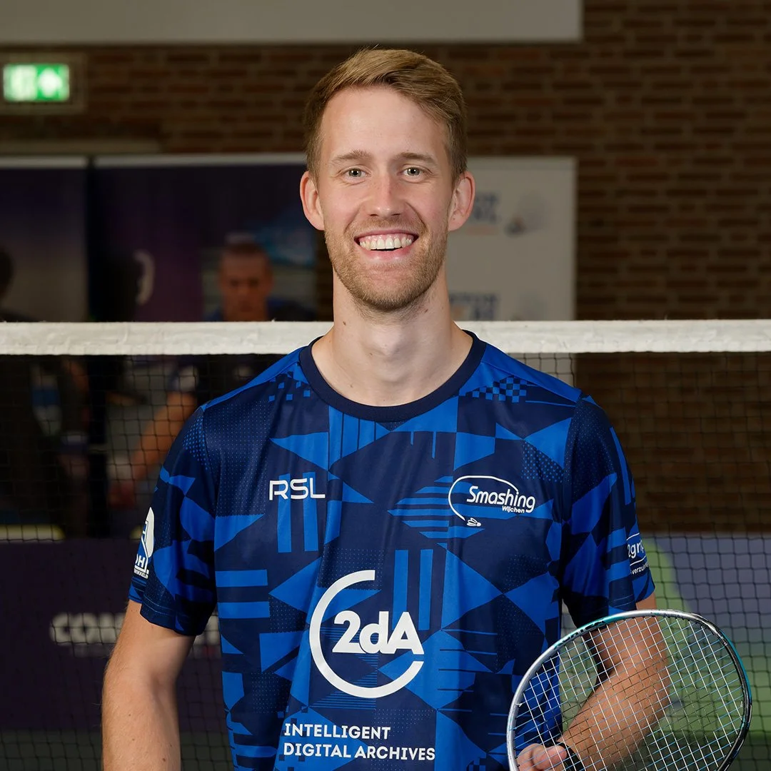 Vrouwelijke badmintonspeler in blauwe sportoutfit met badmintonracket, lachend, in indoor sporthal.