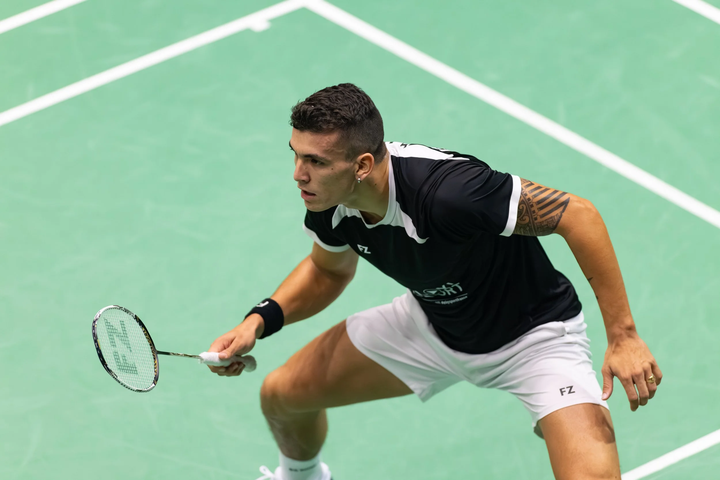 William Villeger speelt badminton op een groene baan, in een lage glimlachende houding met een badmintonracket in de hand.