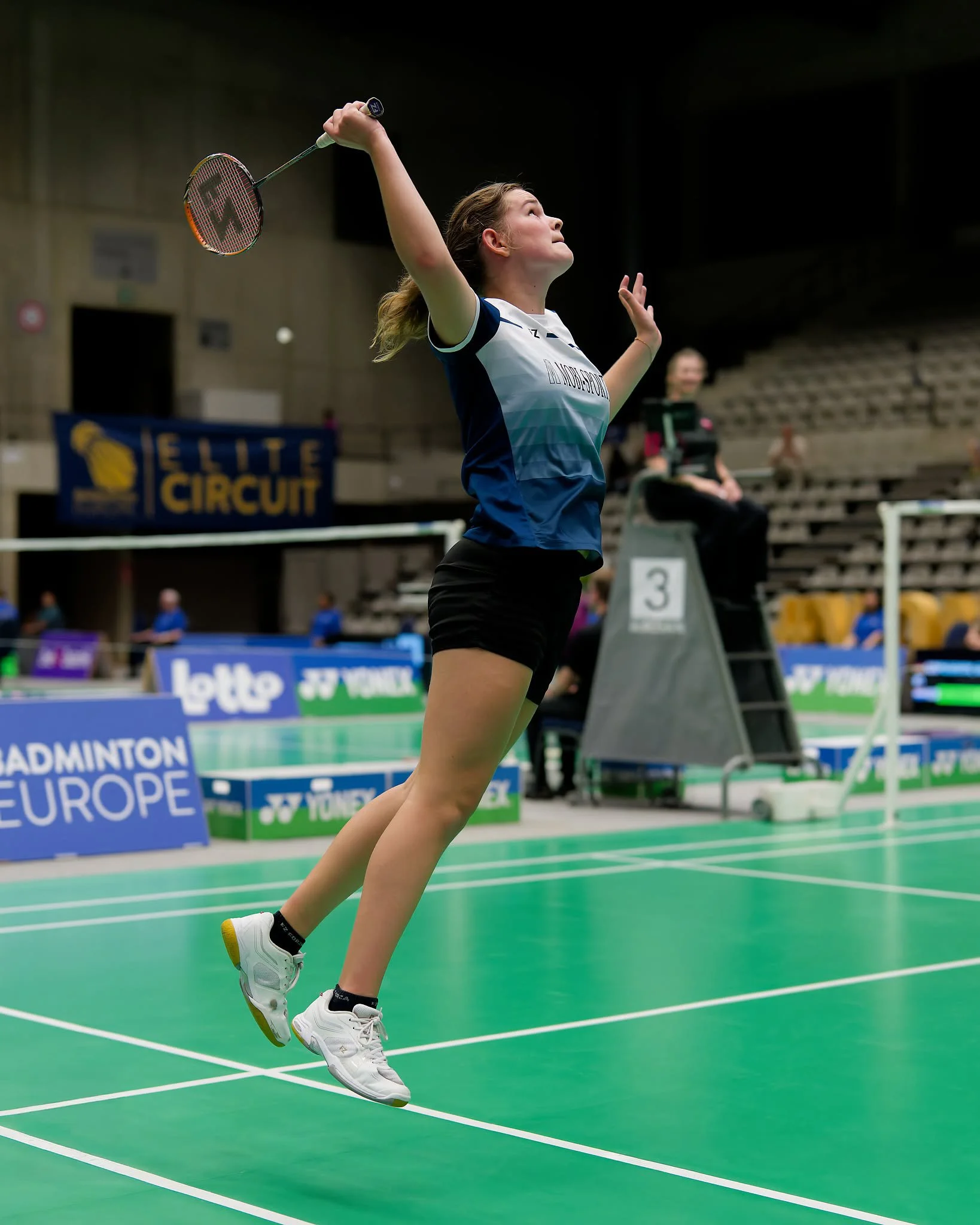 Lieke van Parys springt hoog tijdens een wedstrijd, met haar arm uitgestrekt naar boven, klaar om de shuttle te raken op een indoor badmintonbaan.