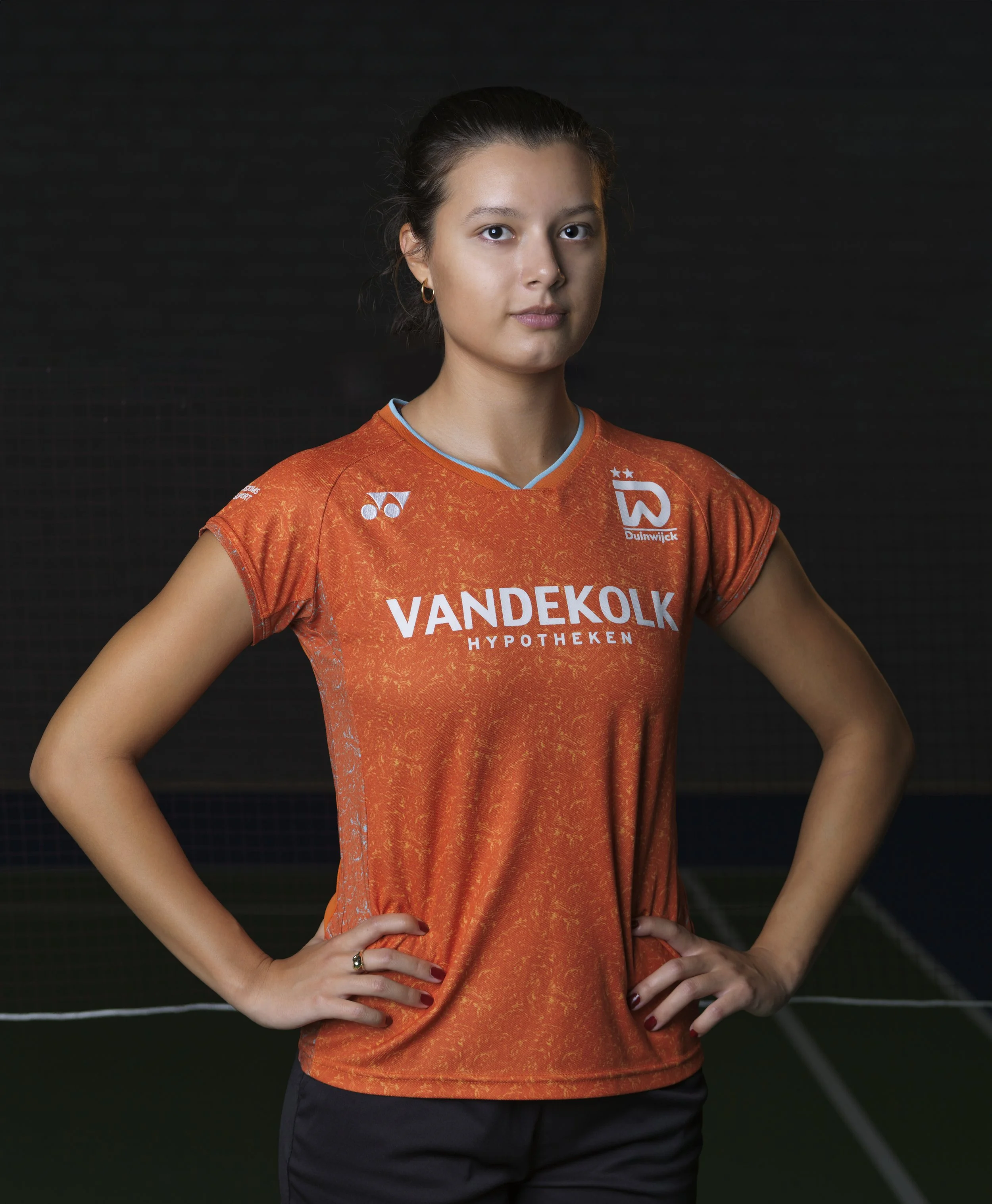 Vrouw in oranje sportshirt met logo's, poseert met handen op heupen, donkere achtergrond.
