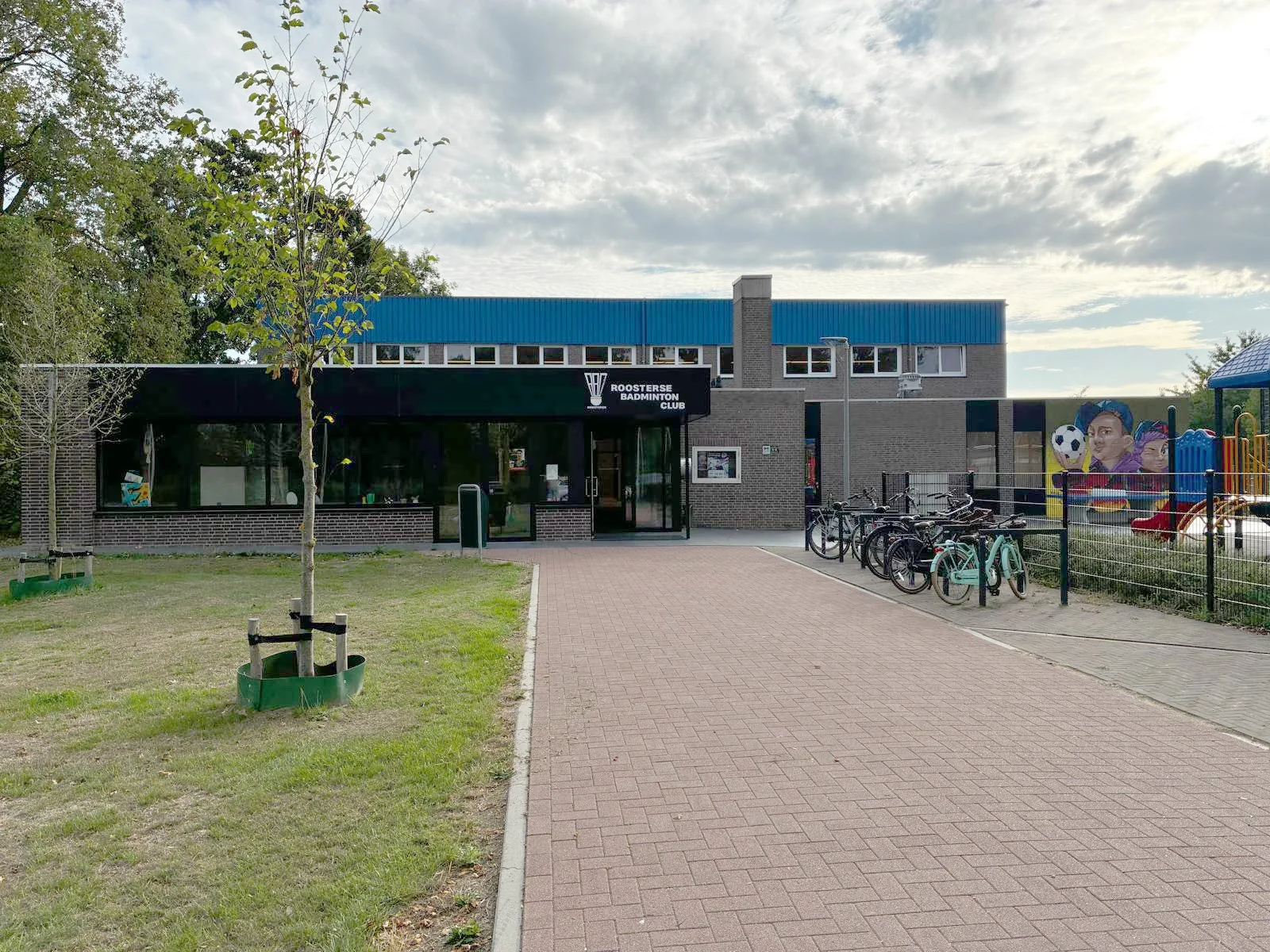 Het gebouw van Roosterse Badminton Club met een parkeerruimte en een kinderspeelplaats ernaast, onder een bewolkte hemel.
