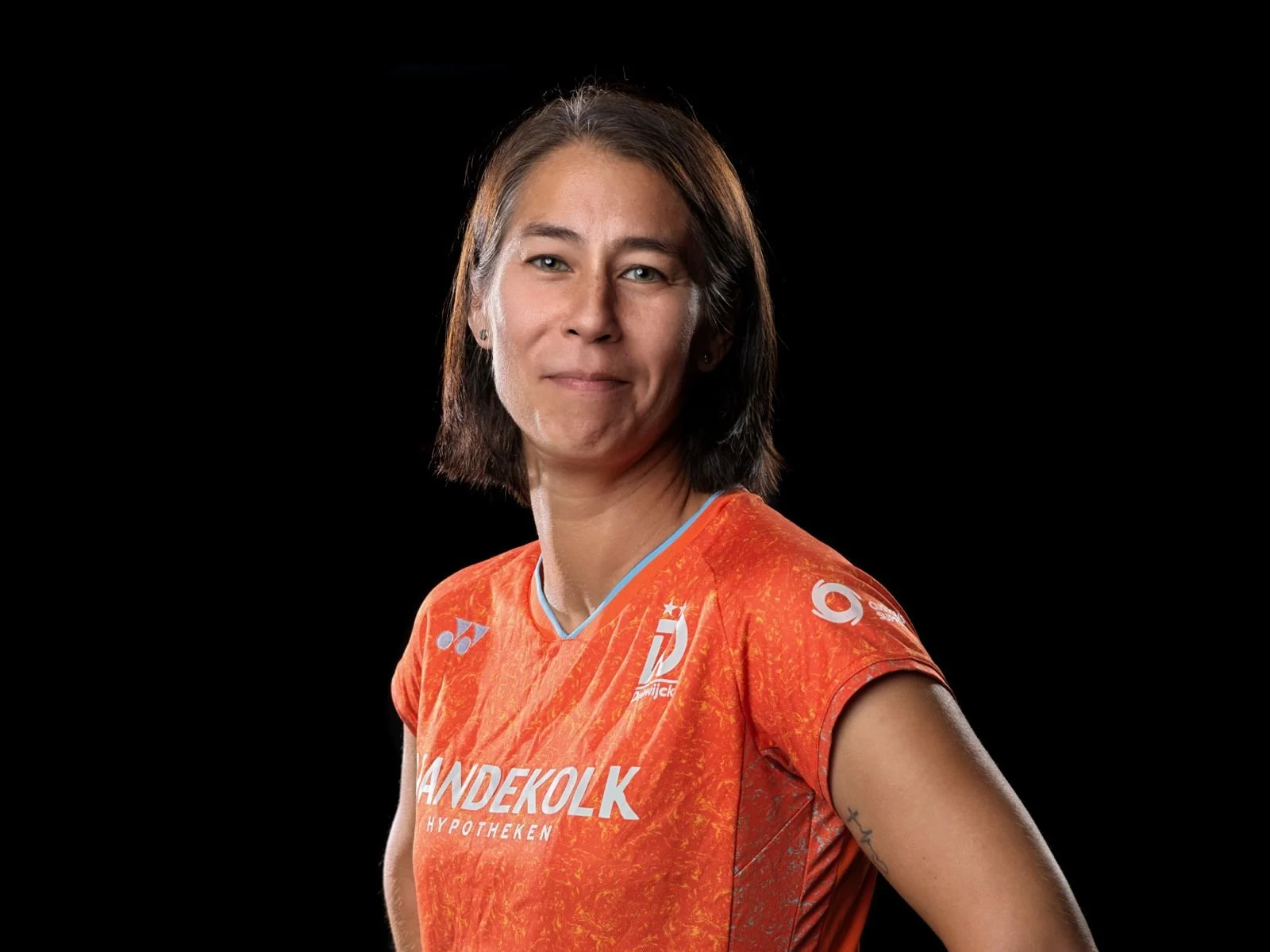 Portret van een vrouw met kort bruin haar, gekleed in een oranje sportshirt met sponsorlogo's, tegen een zwarte achtergrond.