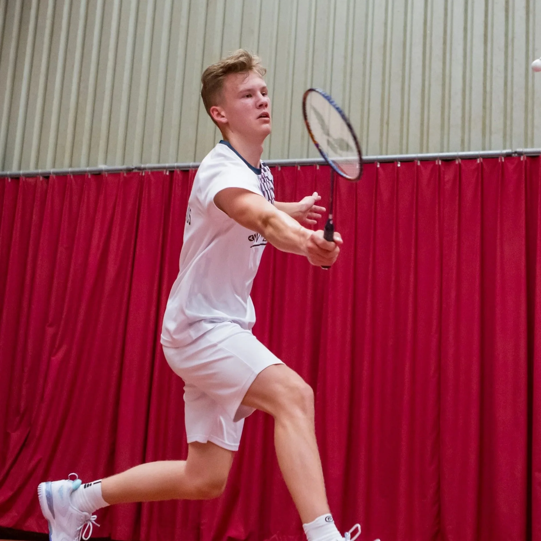 Dion Hoegen die badminton speelt op een indoor baan met rode gordijnen op de achtergrond, hij is gekleed in een wit sportshirt, witte shorts, witte sportschoenen en witte sokken.