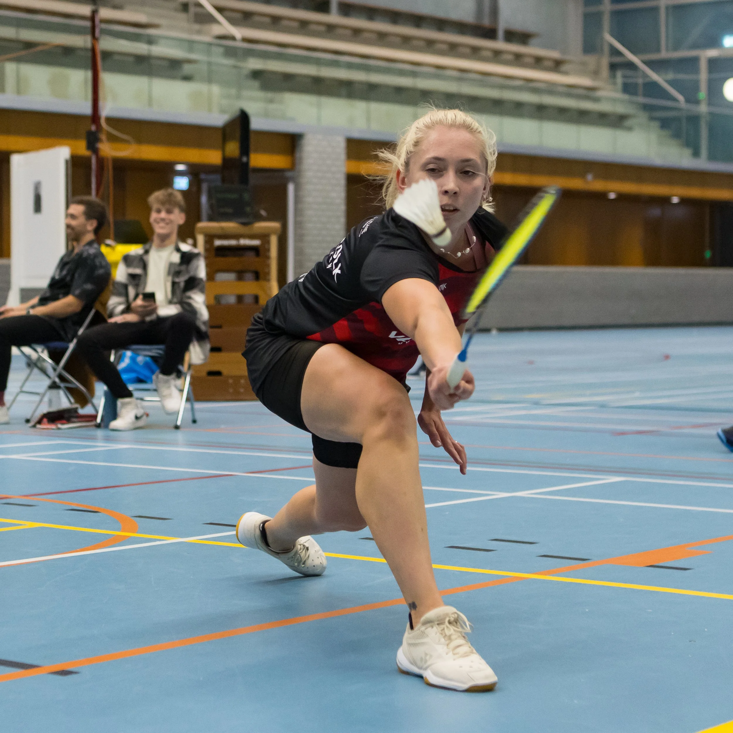 Natalia Perminova die badminton speelt in een indoorsporthal, met twee lachende jongens op de achtergrond die op stoelen zitten.