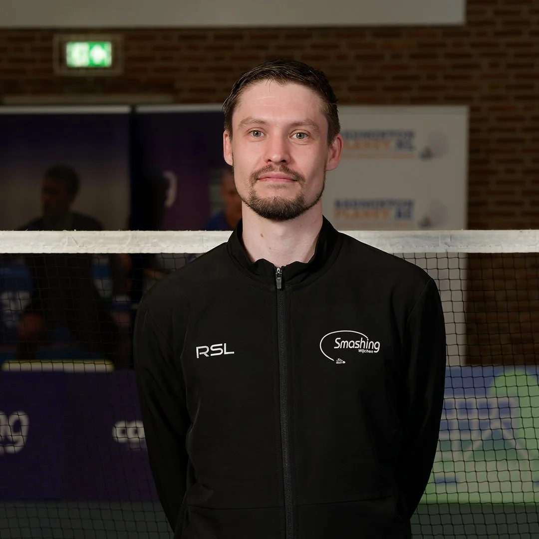 Man poseert voor een net op een indoor tennisbaan, dragend een zwarte sportjas met logo's.