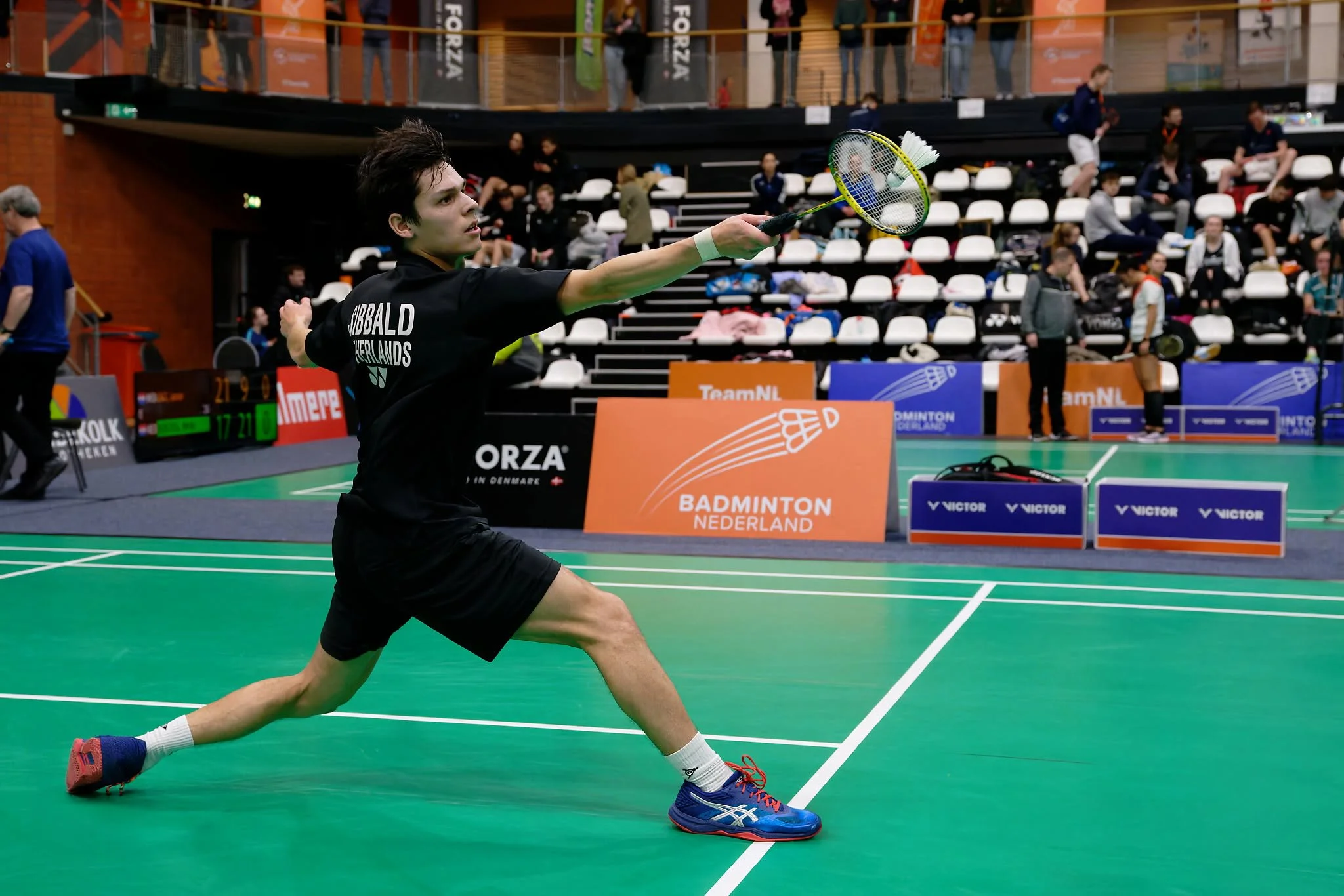 Thomas SIbbald die badminton speelt op een indoorbaan, in een uitvalspositie met zijn racketschouder uitgestrekt, tussen de lijnen van de groene banen.