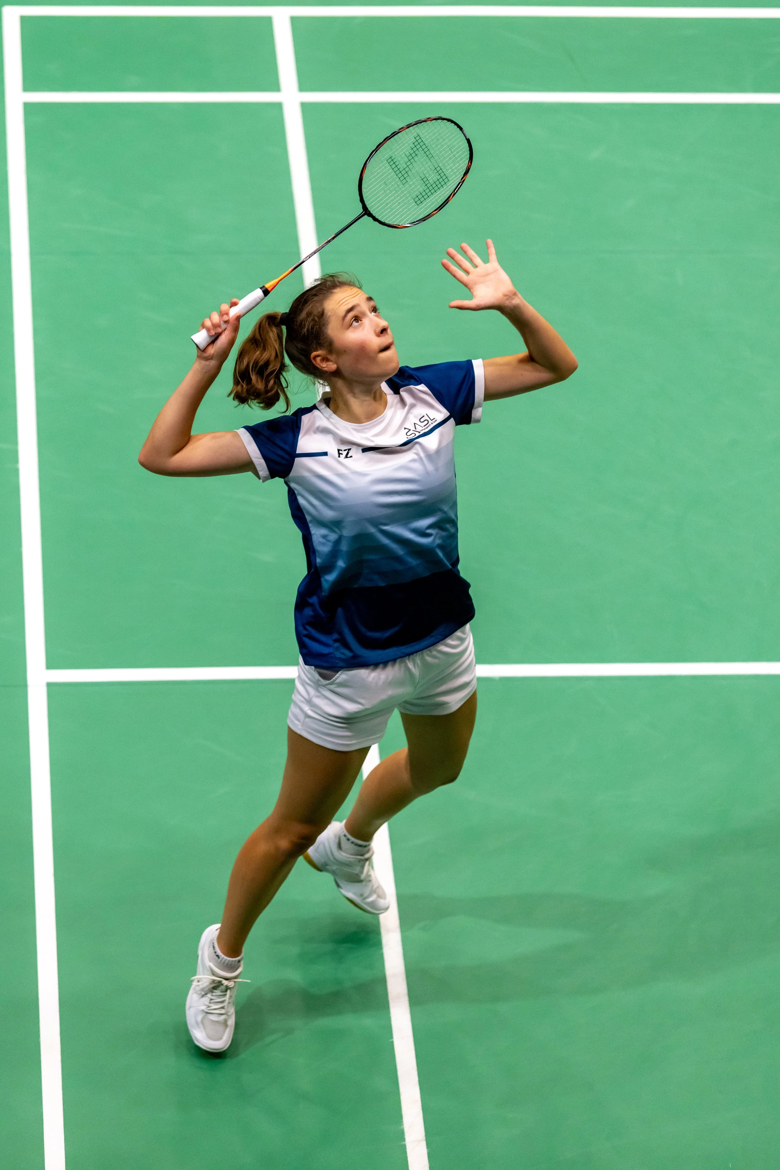 Lien Lammertyn die badminton speelt op groen indoor sportveld, zwaai met haar racket en lijkt klaar om te slaan.