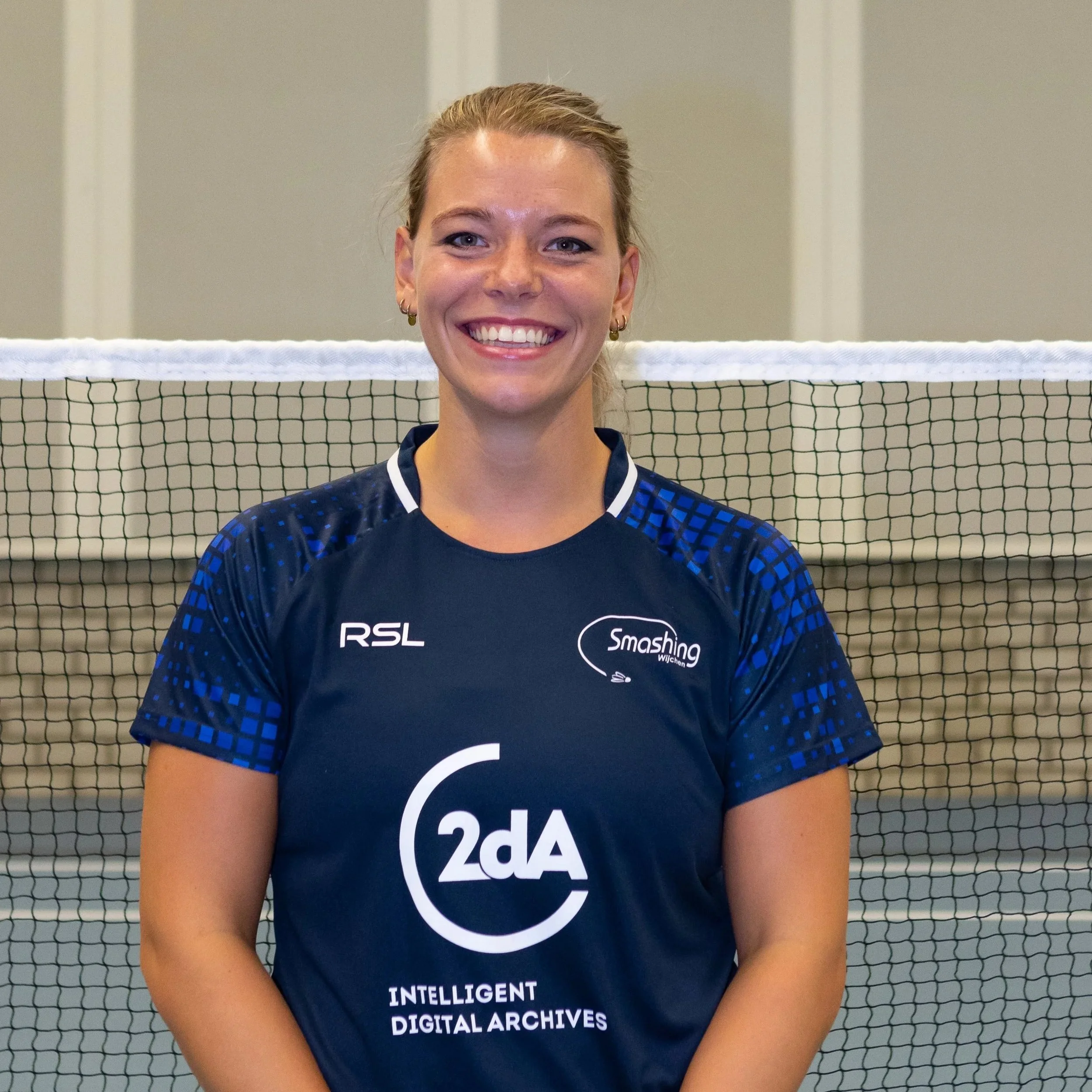Lieke Maes als glimlachende jonge vrouw met blond haar voor een badminton net, sportkleding met logo's, binnen.